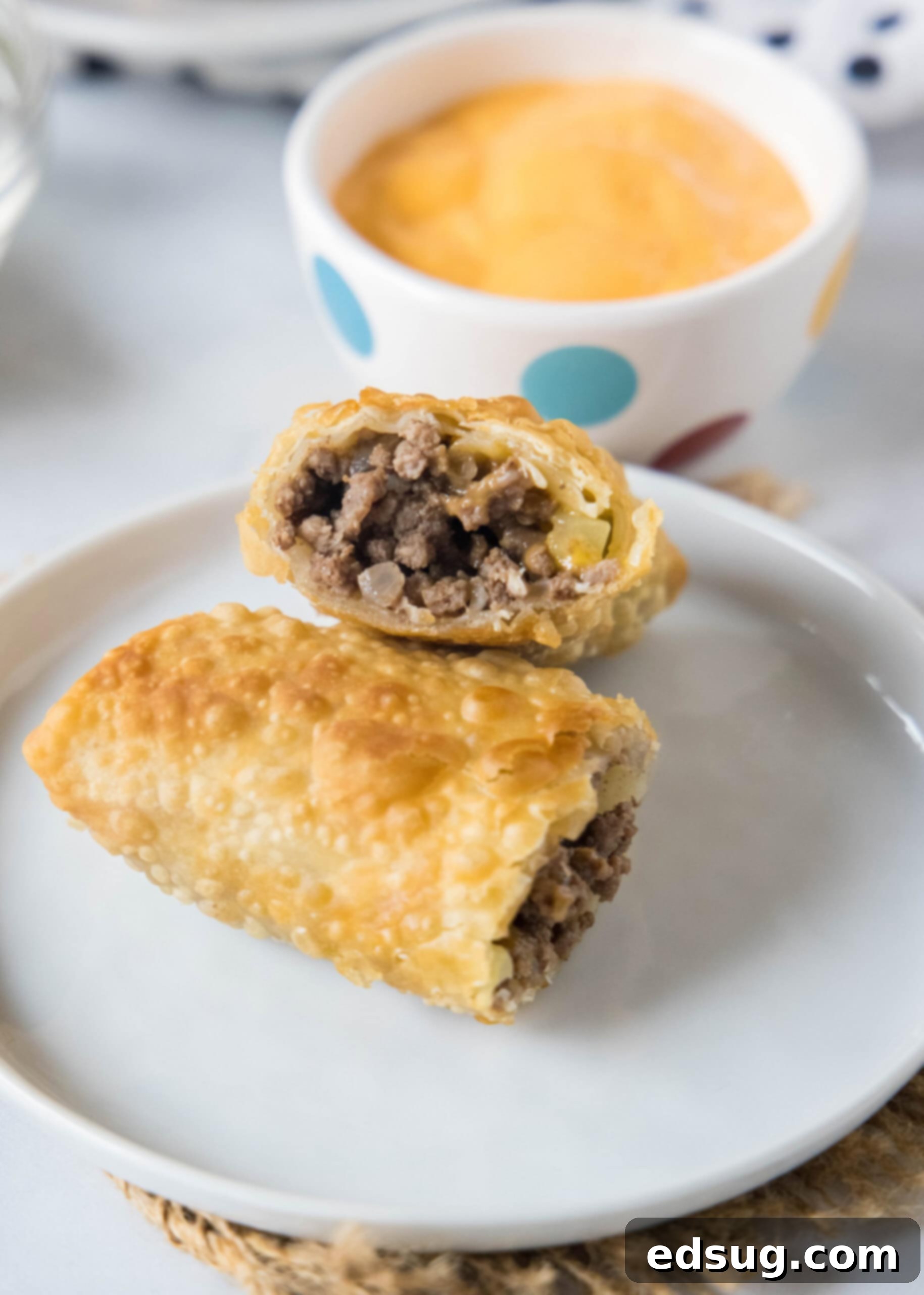 Looking for a fun and easy appetizer recipe? Try these cheeseburger egg rolls complete with creamy, tangy homemade burger sauce for dipping! Two halves of a cheeseburger egg roll stacked on a white plate with a bowl of dipping sauce in the background.