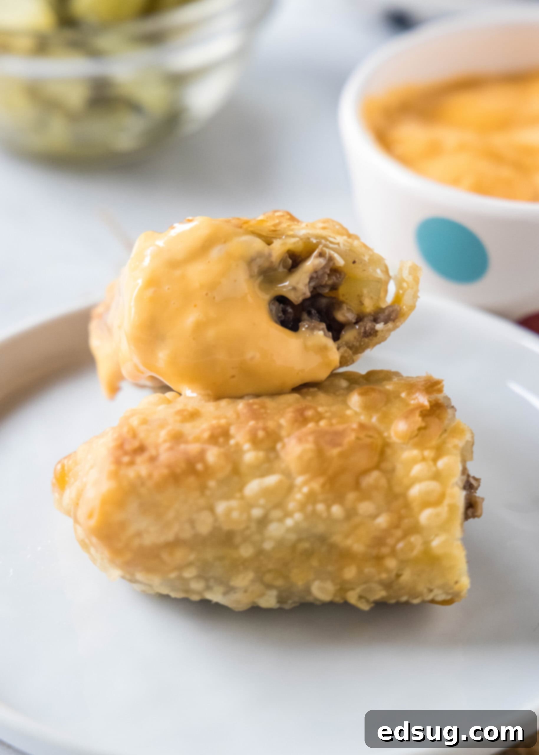 Looking for a fun and easy appetizer recipe? Try these cheeseburger egg rolls complete with creamy, tangy homemade burger sauce for dipping! Two halves of a cheeseburger egg roll stacked on a white plate with a bowl of dipping sauce in the background.