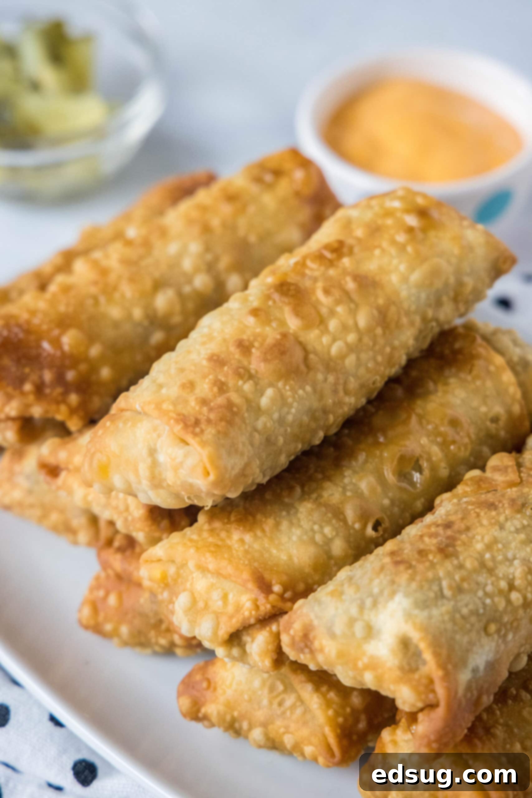 Looking for a fun and easy appetizer recipe? Try these cheeseburger egg rolls complete with creamy, tangy homemade burger sauce for dipping! Cheeseburger egg rolls stacked on a platter with a bowl of dipping sauce in the background.