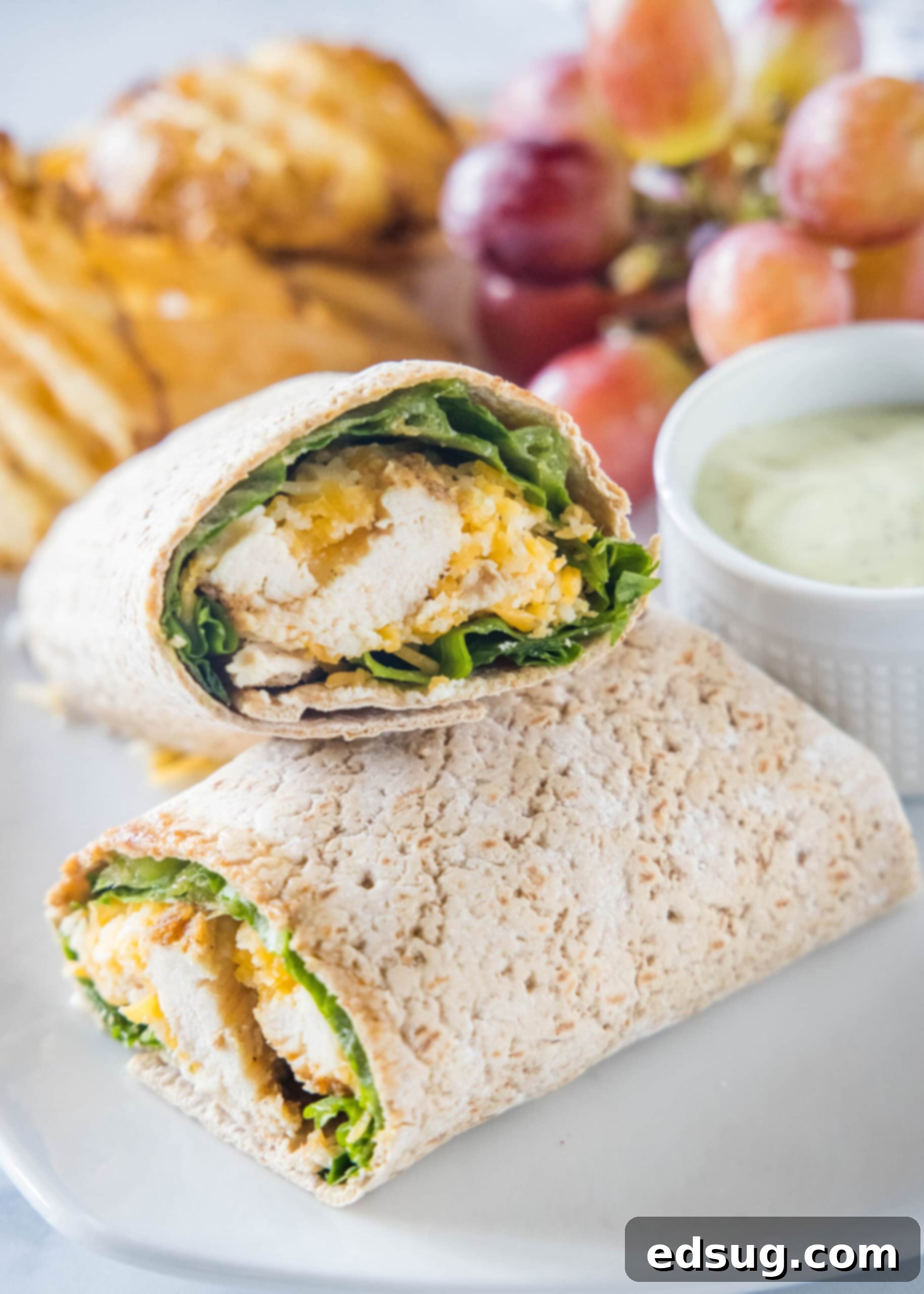 This easy Chick Fil A Cool Wrap recipe is the best excuse to skip the drive-thru! Enjoy this copycat at home with your favorite dipping sauce. Close up of two halves of a Chick Fil A Cool Wrap on a plate next to a bowl of dipping sauce, grapes, and potato chips.