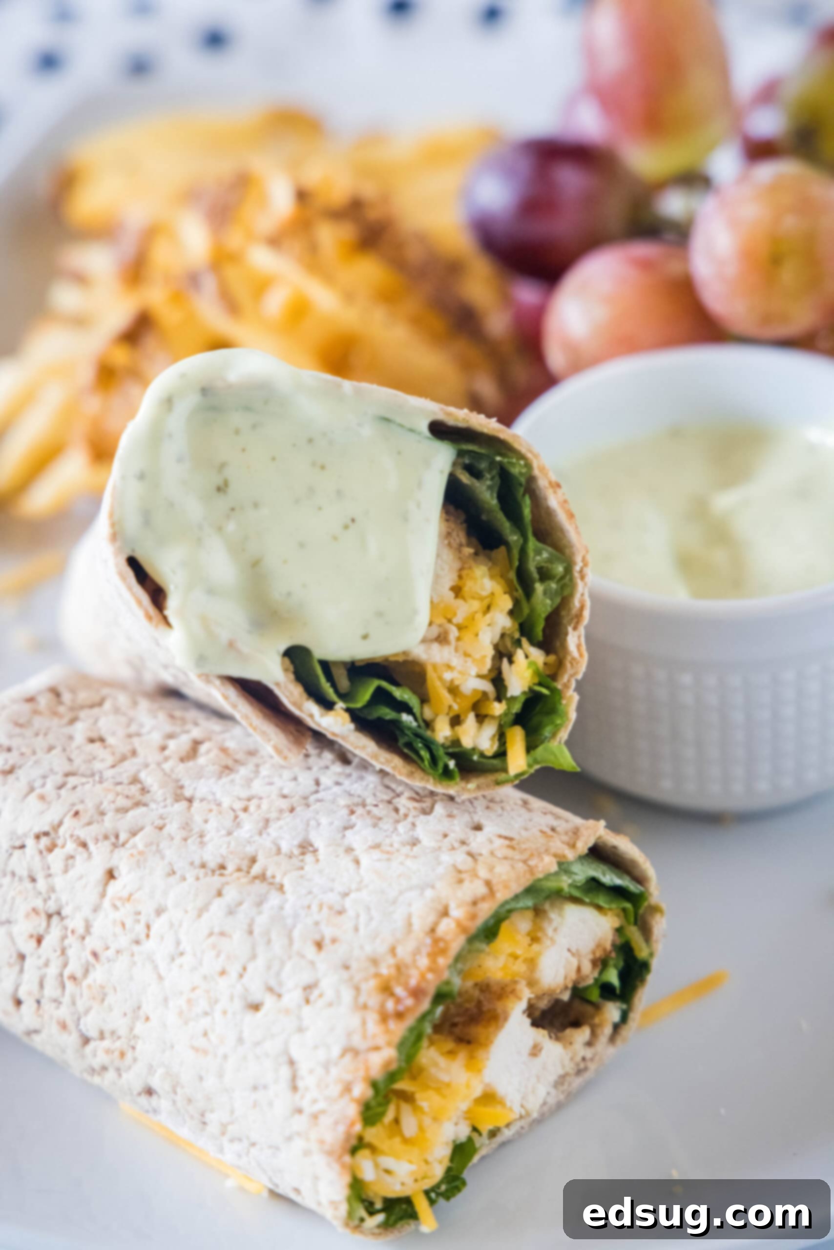 This easy Chick Fil A Cool Wrap recipe is the best excuse to skip the drive-thru! Enjoy this copycat at home with your favorite dipping sauce. Two halves of a Chick Fil A Cool Wrap on a plate, with one half covered in dipping sauce, with a bowl of dipping sauce, grapes, and potato chips in the background.