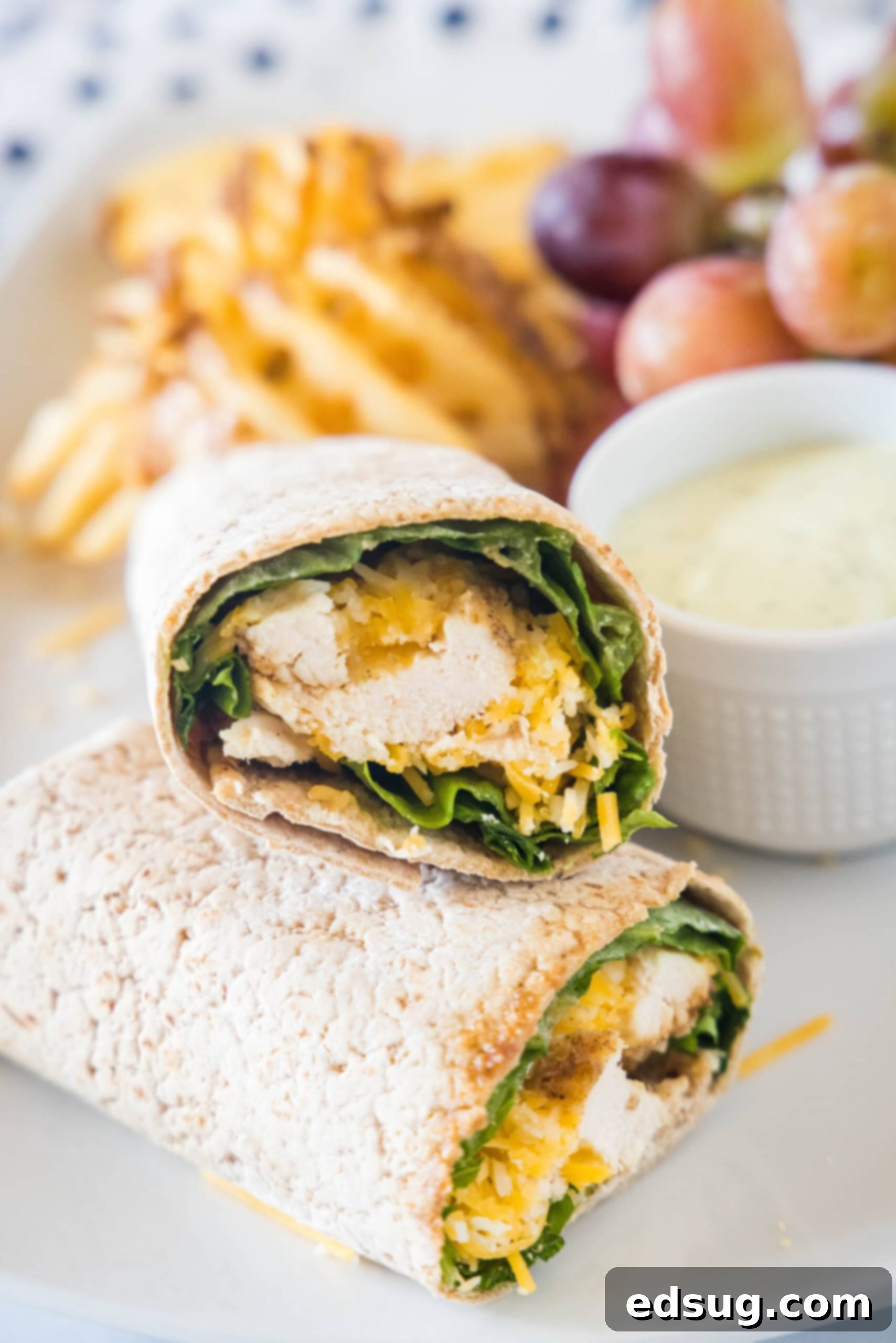 This easy Chick Fil A Cool Wrap recipe is the best excuse to skip the drive-thru! Enjoy this copycat at home with your favorite dipping sauce. Two halves of a Chick Fil A Cool Wrap on a plate next to a bowl of dipping sauce, grapes, and potato chips.