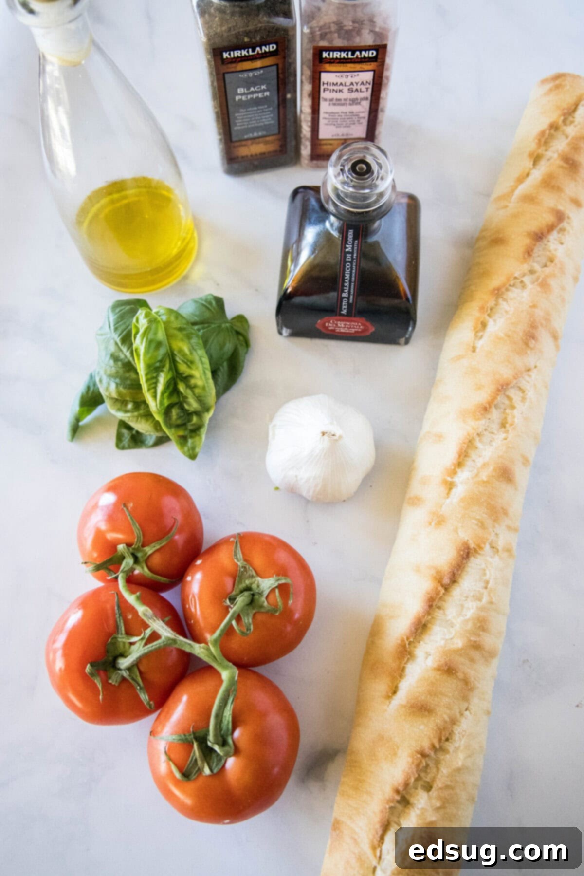 This easy bruschetta is the perfect appetizer or party food. It's loaded with tomatoes, garlic, and basil, and only takes 5 minutes. Overhead view of the ingredients needed for bruschetta: A baguette, four tomatoes on the vine, a head of garlic, a bunch of basil, a jar of balsamic vinegar, a bottle of olive oil, and salt and pepper