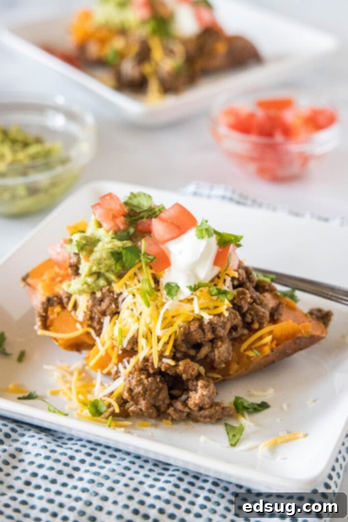 These taco stuffed sweet potatoes are soft and fluffy sweet potatoes piled high with savory ground beef and your favorite taco toppings! A taco sweet potato piled with taco toppings served on a white plate, with a second sweet potato and bowls of toppings in the background.