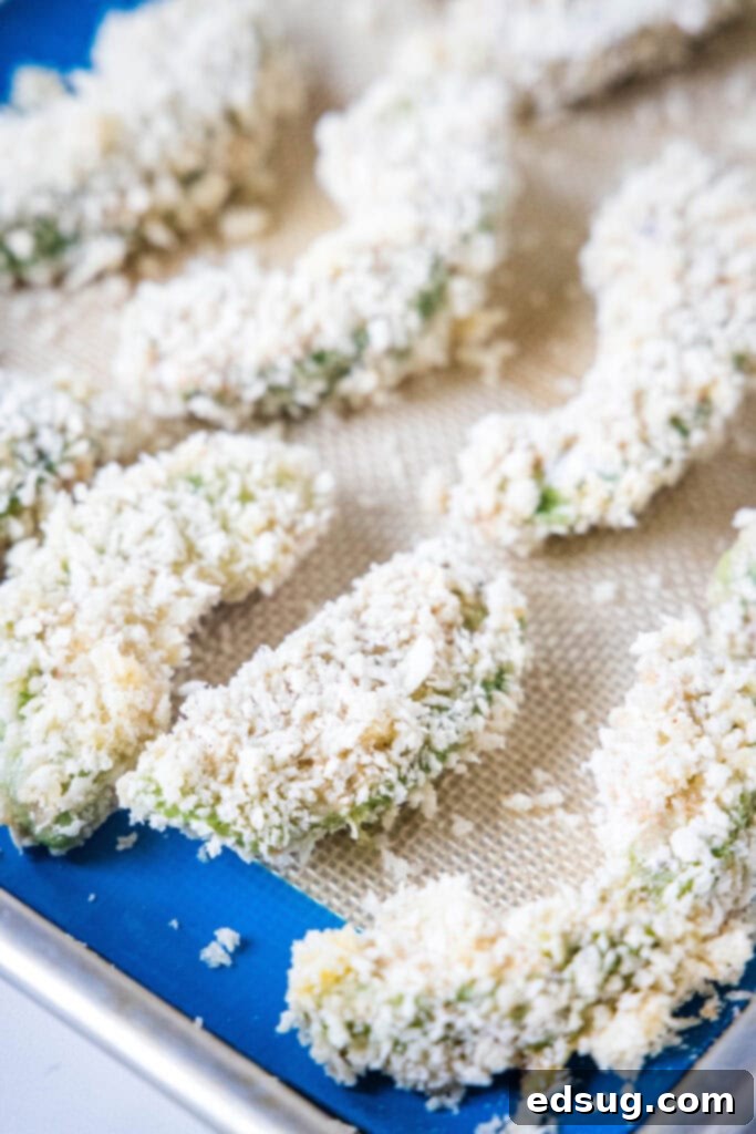Avocado fries are crispy on the outside and warm and creamy on the inside, made in the air fryer (or oven). Don't forget the dipping sauce! Breaded avocado slices neatly arranged on a Silpat-lined baking sheet, prepared for cooking.