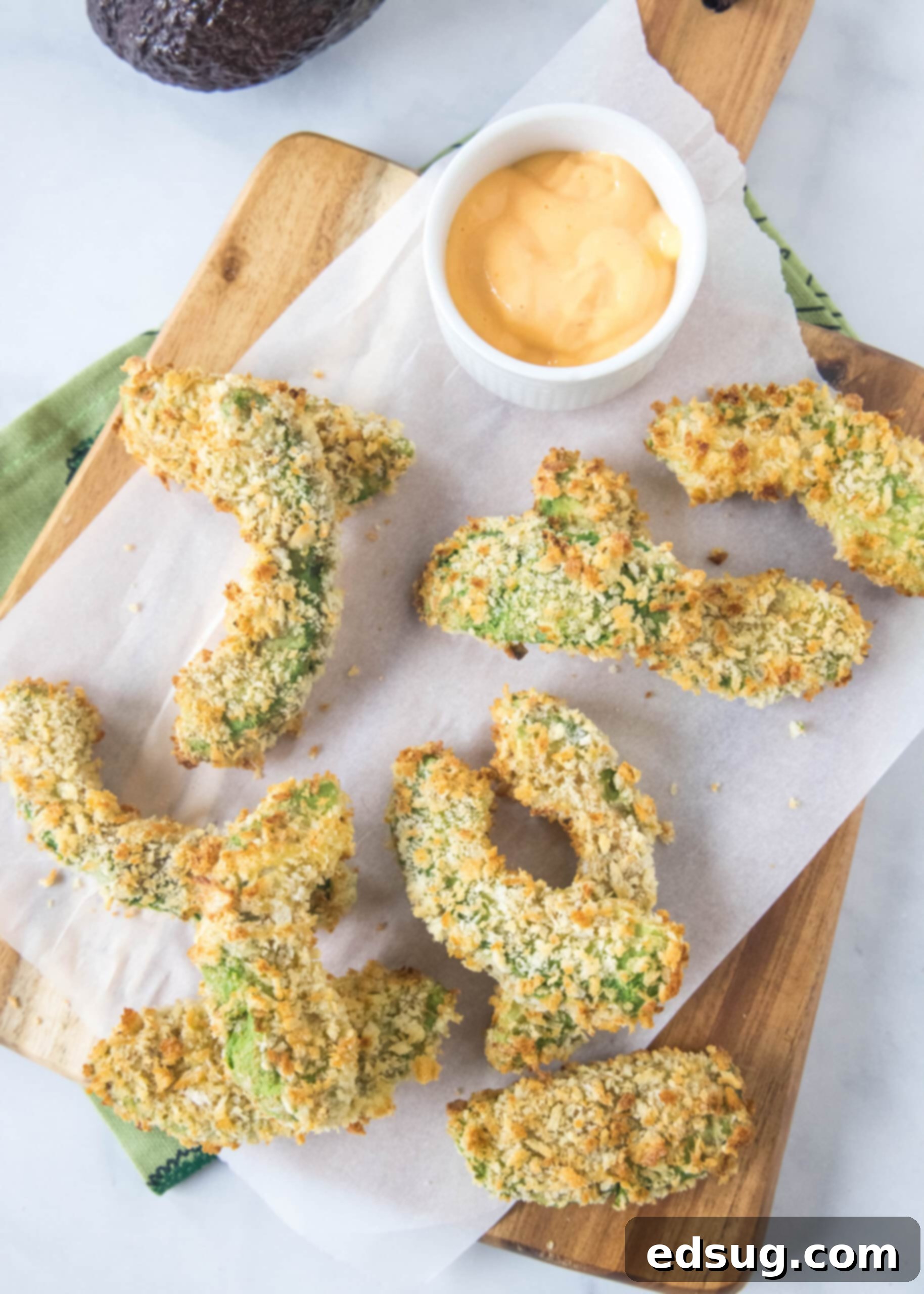 Avocado fries are crispy on the outside and warm and creamy on the inside, made in the air fryer (or oven). Don't forget the dipping sauce! Overhead view of crispy avocado fries scattered on a wooden cutting board with parchment paper, next to a small bowl of dipping sauce, ready to be enjoyed.