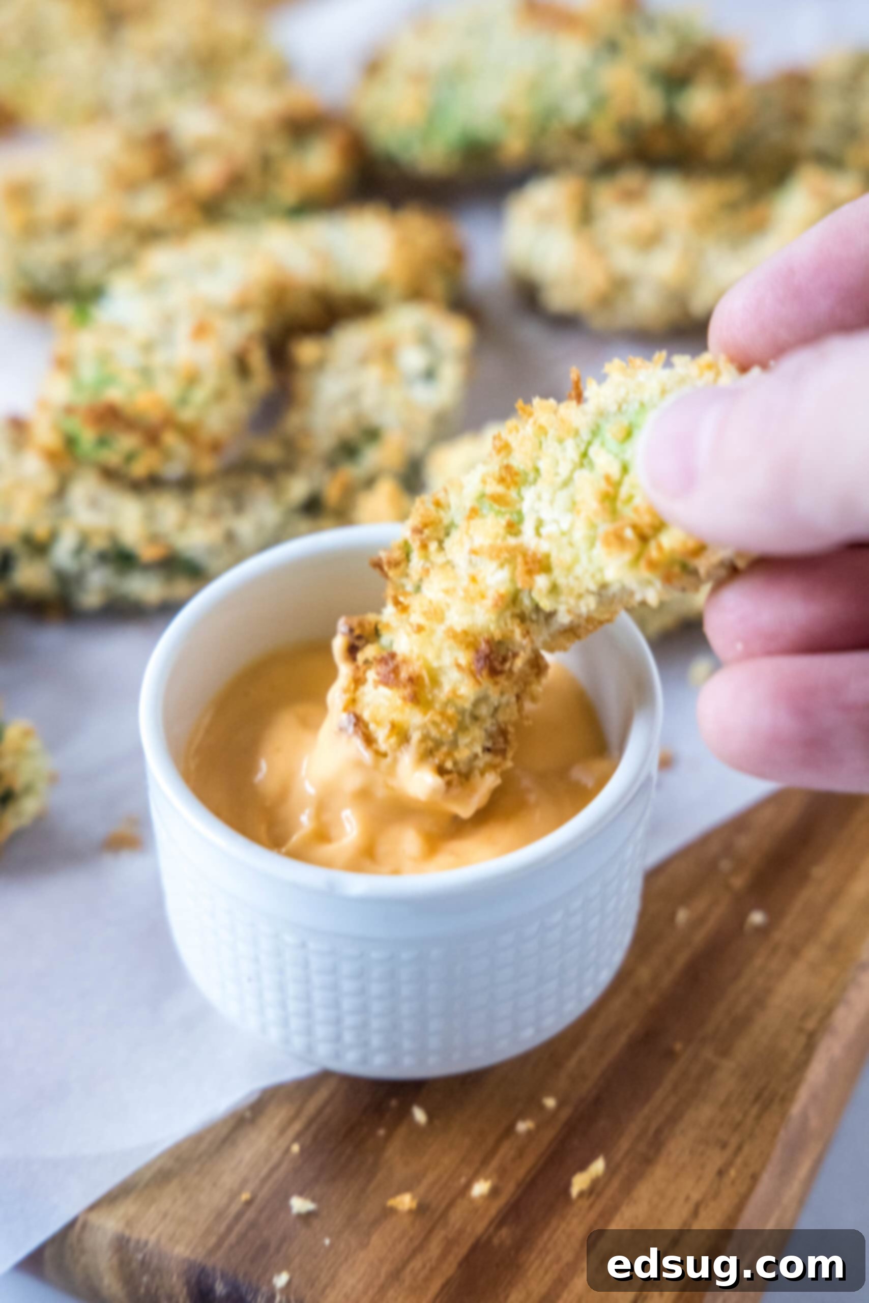 Avocado fries are crispy on the outside and warm and creamy on the inside, made in the air fryer (or oven). Don't forget the dipping sauce! A hand dipping a golden, crispy avocado fry into a small bowl of creamy dipping sauce, with more fries blurred in the background.