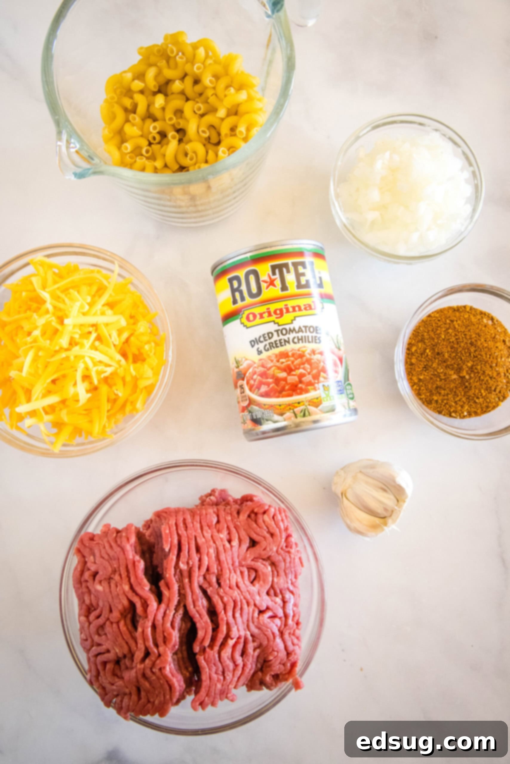 A collection of fresh ingredients laid out on a wooden board for making taco mac and cheese, including ground beef, pasta, cheese, onions, and tomatoes.