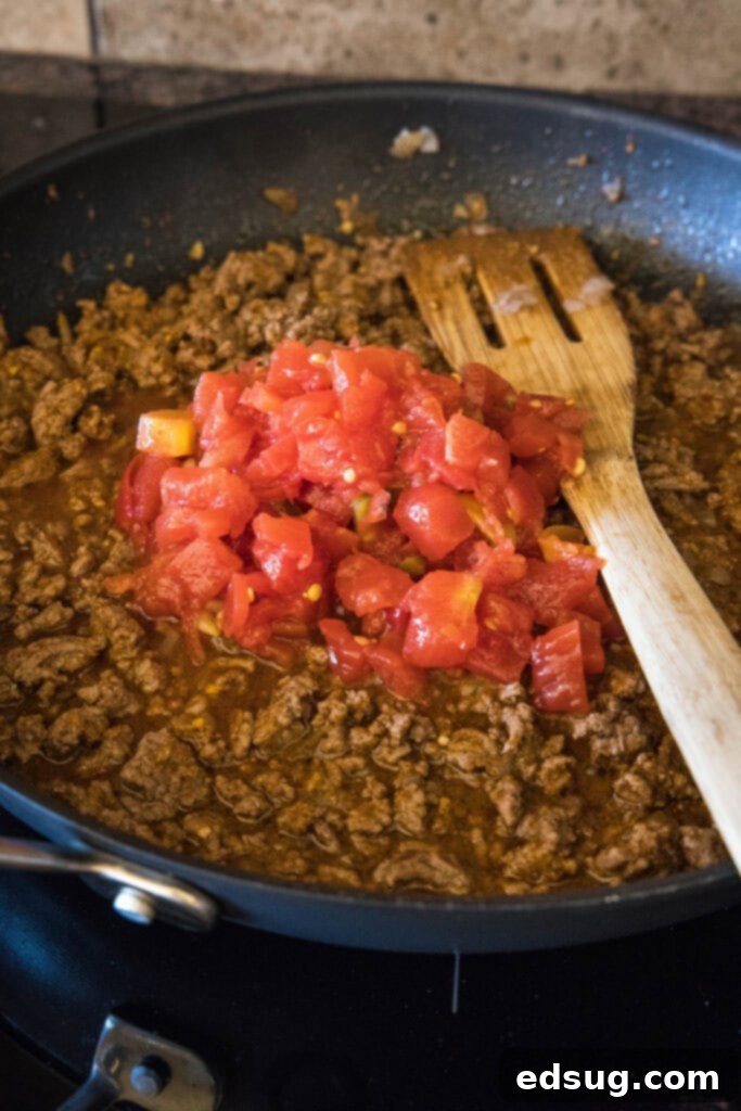 Canned diced tomatoes and taco seasoning are added to a skillet with browned ground beef and onions, creating a rich sauce.