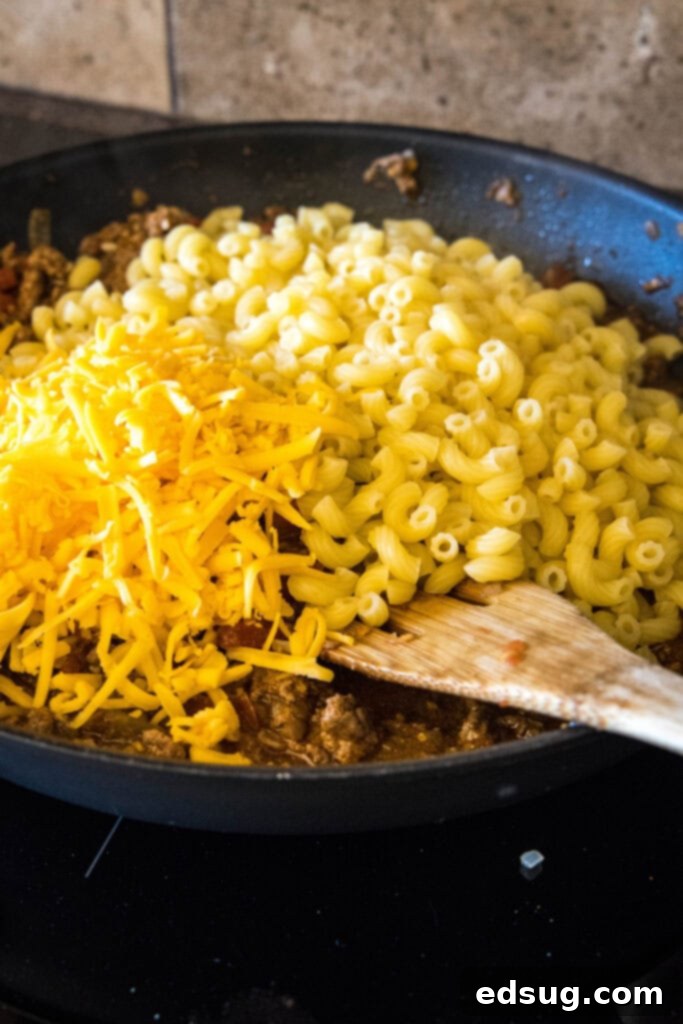 Cooked elbow macaroni noodles and shredded cheddar cheese are stirred into the skillet with taco meat and tomato sauce, creating a cheesy, saucy mixture.