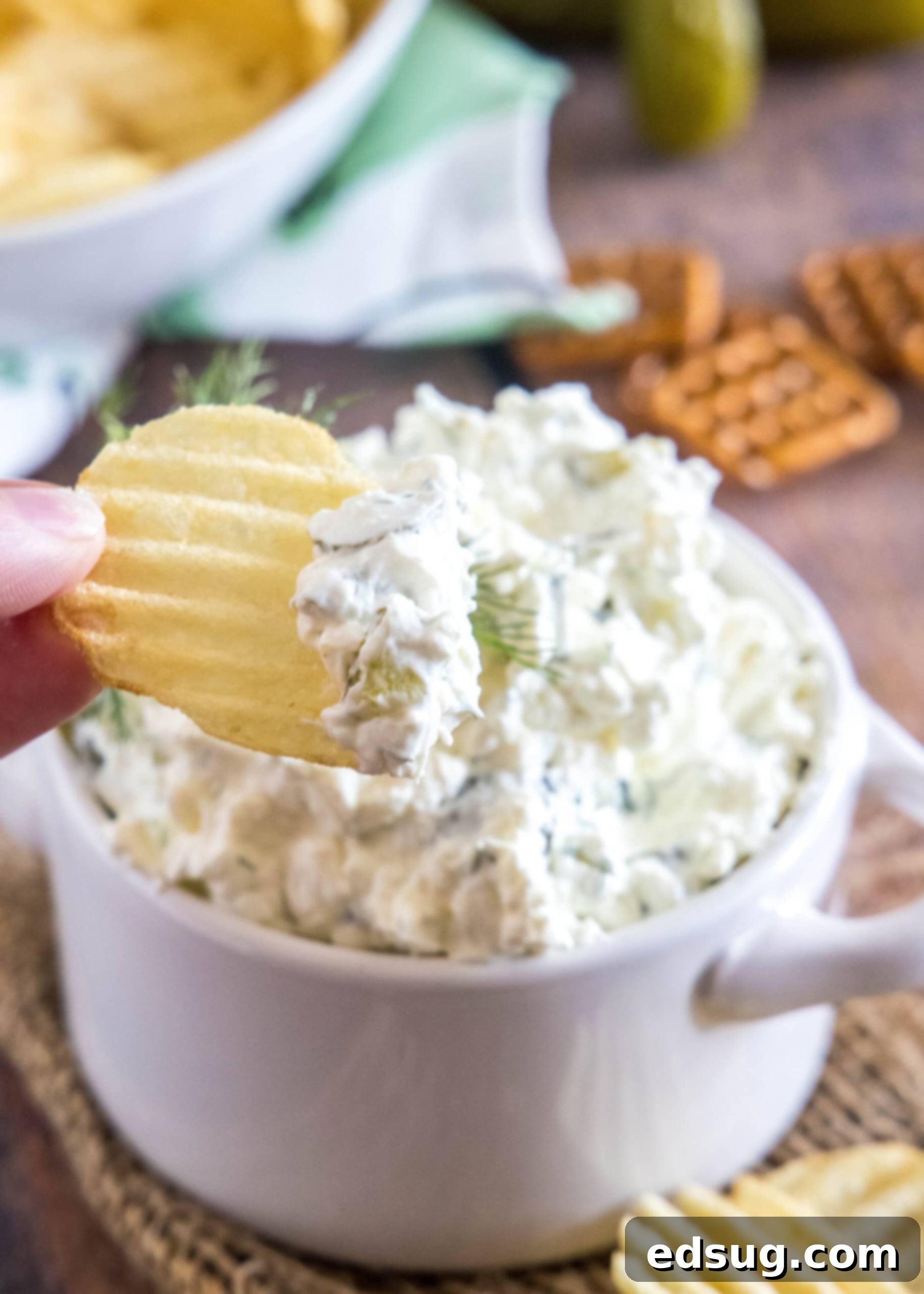 Creamy dill pickle dip is briny, cool, and crunchy. Made with 5 ingredients in minutes, it's delicious for dipping just about everything! Close-up of a hand dipping a ruffled potato chip into creamy dill pickle dip, with a full bowl of dip in the background.