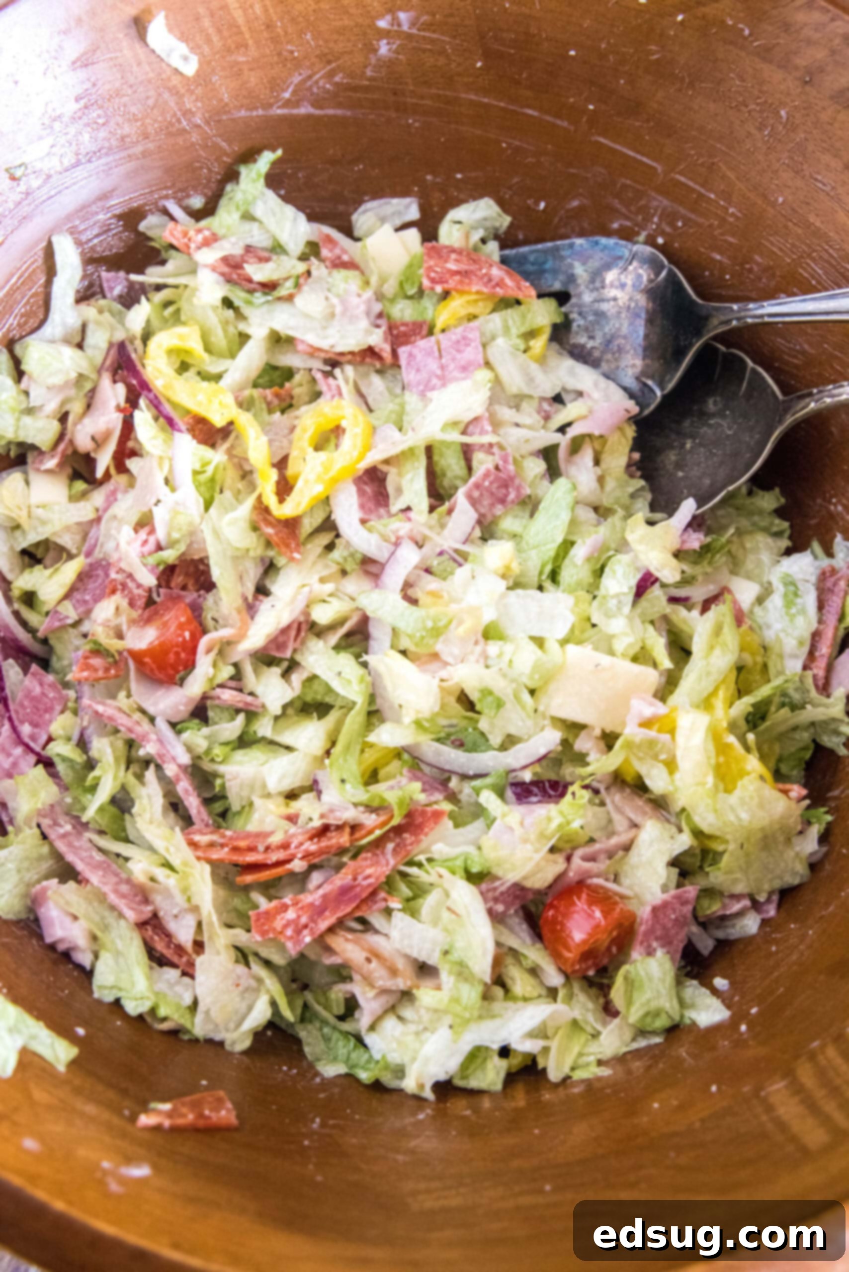 Grinder salad fully tossed with dressing in a rustic wooden bowl, with salad tongs resting on the side, ready for serving.