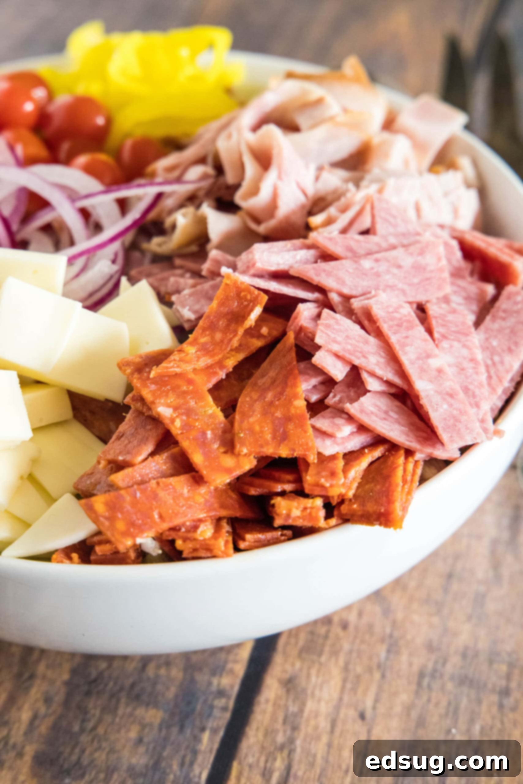 A large ceramic bowl filled with an un-dressed grinder salad, showcasing the colorful layers of chopped lettuce, deli meats, cheese, tomatoes, and red onion.
