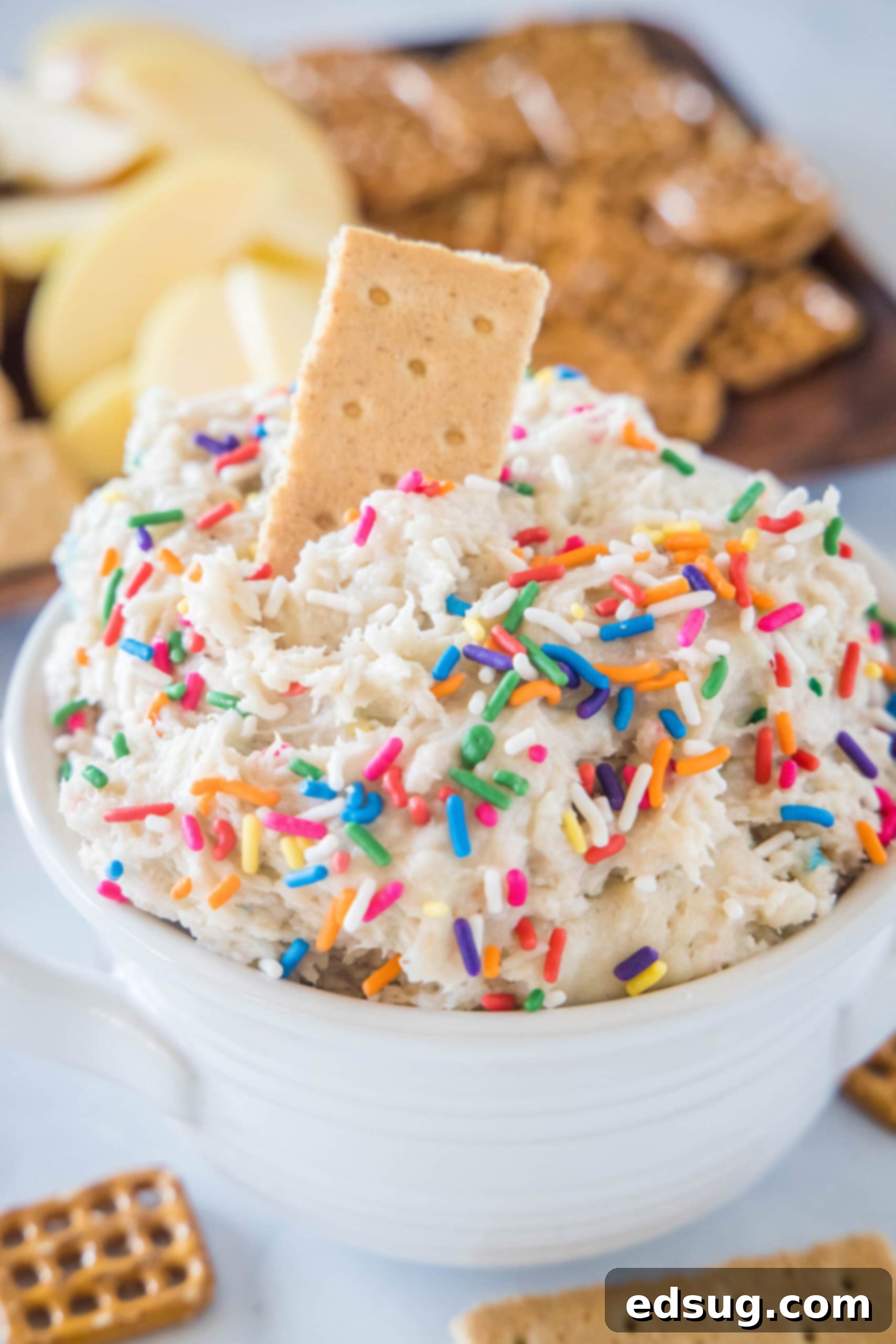 Fluffy funfetti cake batter dip is an easy no-bake dessert made from cake mix and brimming with sprinkles. It tastes just like classic funfetti cake batter! Cake batter dip in a white bowl with a cookie stuck into the top, and a platter of cookies in the background, adorned with colorful sprinkles.