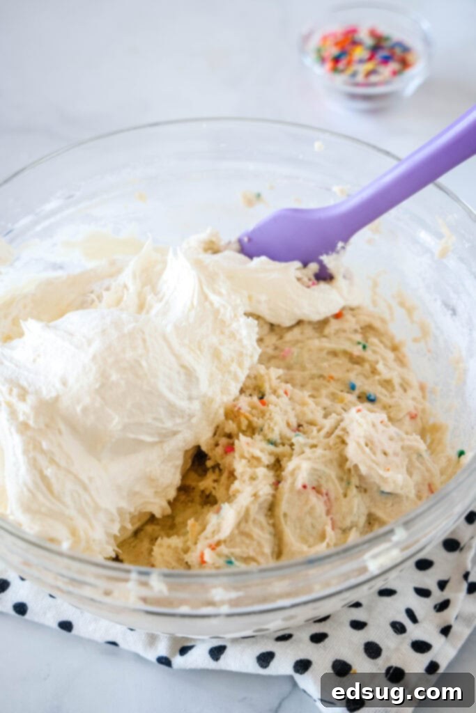 Cool whip partially folded into cake batter dip ingredients in a glass bowl. Cool whip partially folded into cake batter dip ingredients in a glass bowl with a purple rubber spatula.