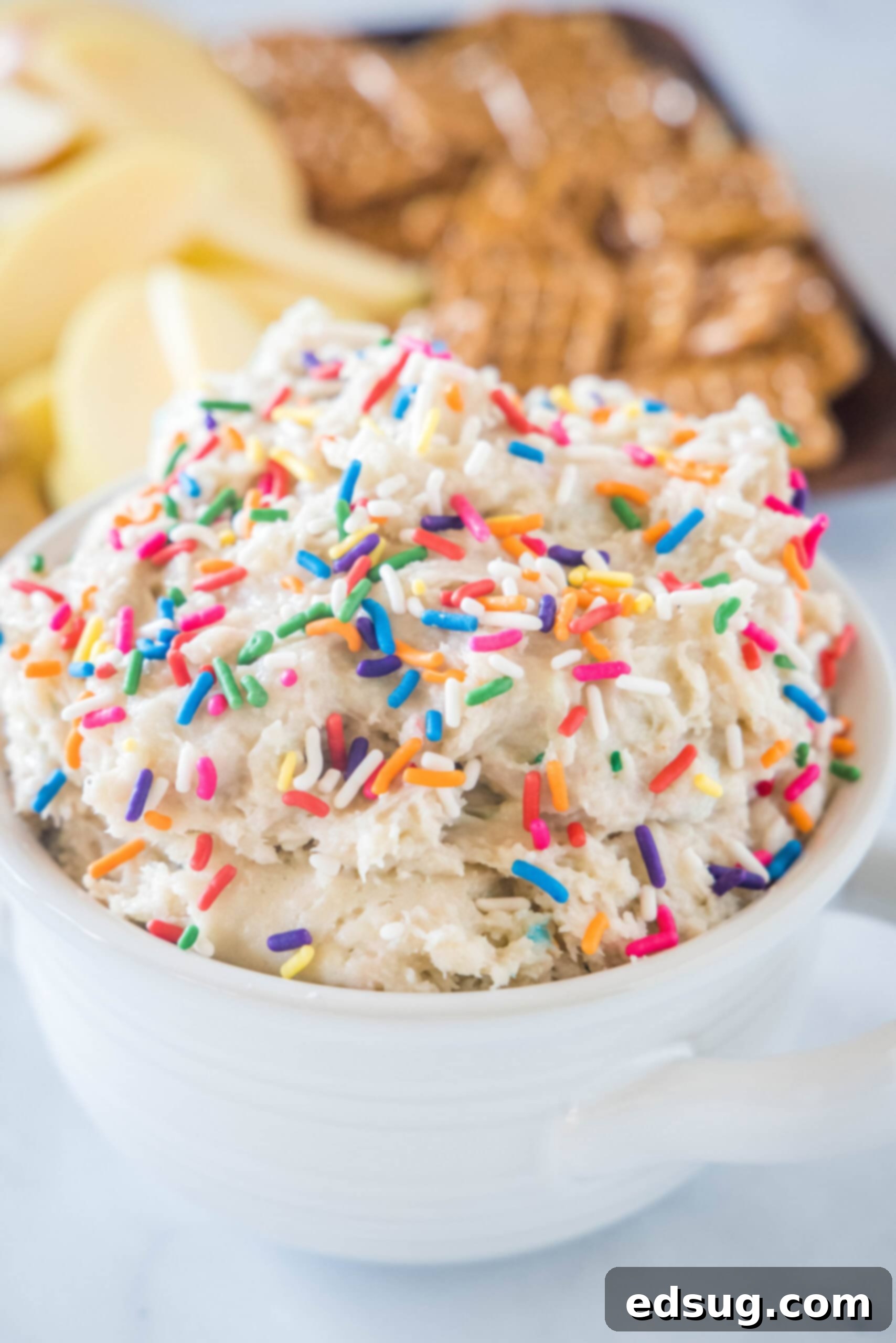 Cake batter dip in a white bowl topped with sprinkles, with a platter of cookies in the background. Cake batter dip in a white bowl topped with sprinkles, with a platter of cookies in the background.