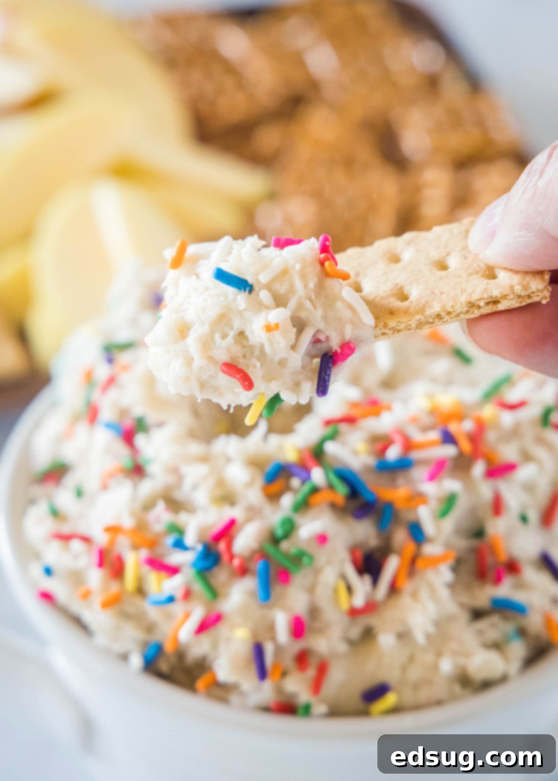 A hand holding a cookie over a bowl of cake batter dip. A hand holding a cookie over a bowl of cake batter dip, with a platter of cookies in the background.