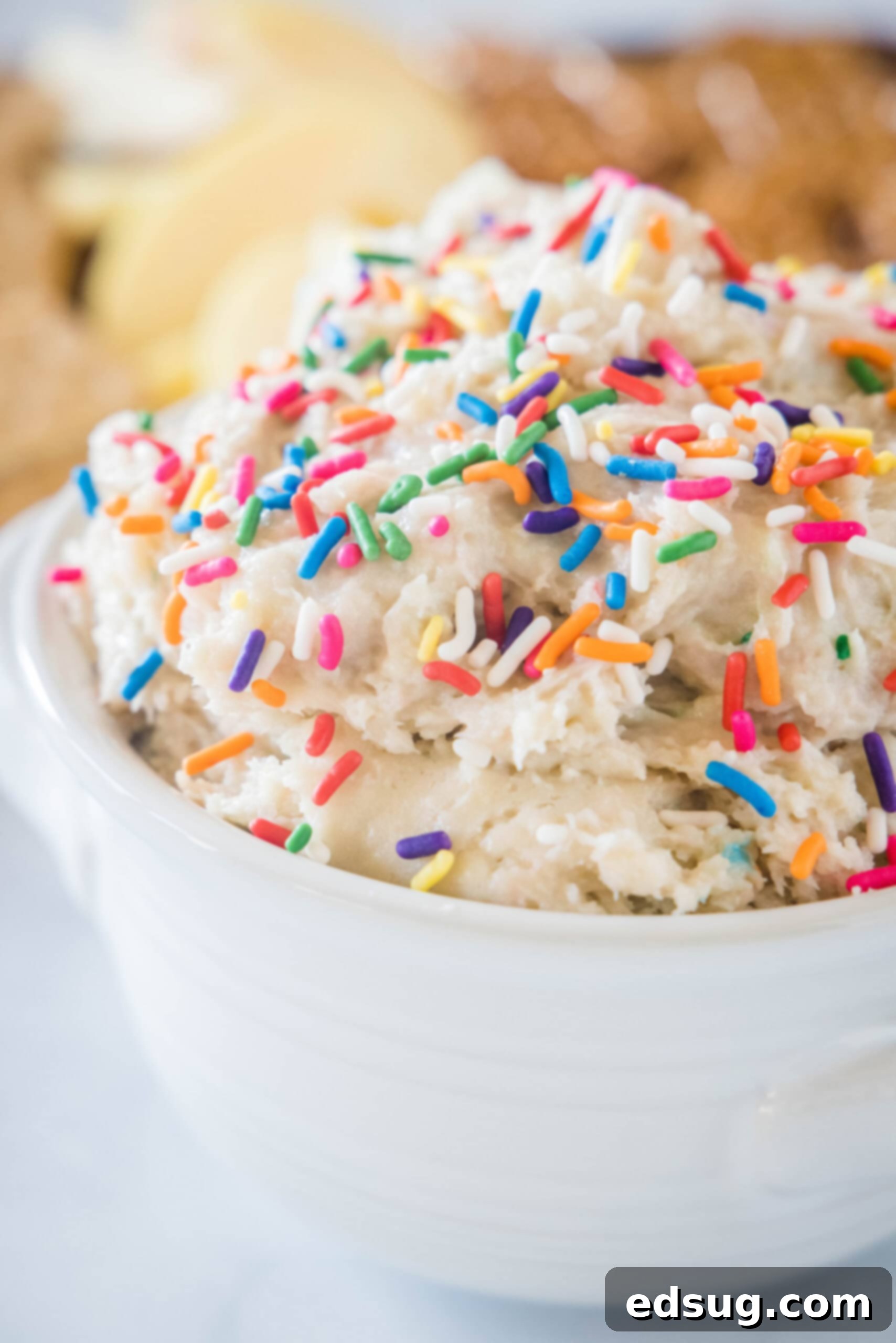 Cake batter dip in a white bowl topped with sprinkles. Cake batter dip in a white bowl topped with sprinkles, with a platter of cookies in the background.