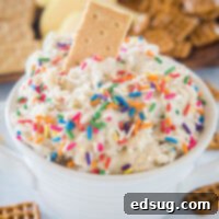 Cake batter dip in a white bowl with a cookie stuck into the top, and a platter of cookies in the background.