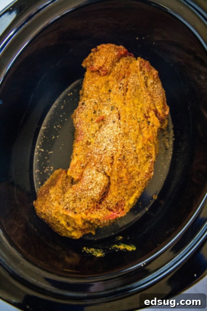 Seasoned chuck roast coated with mustard and dry rub placed inside the crock pot, ready to cook.