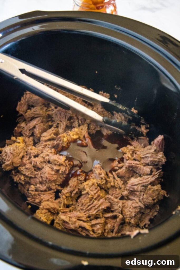 Metal tongs resting in a mound of perfectly shredded BBQ beef inside the crock pot.