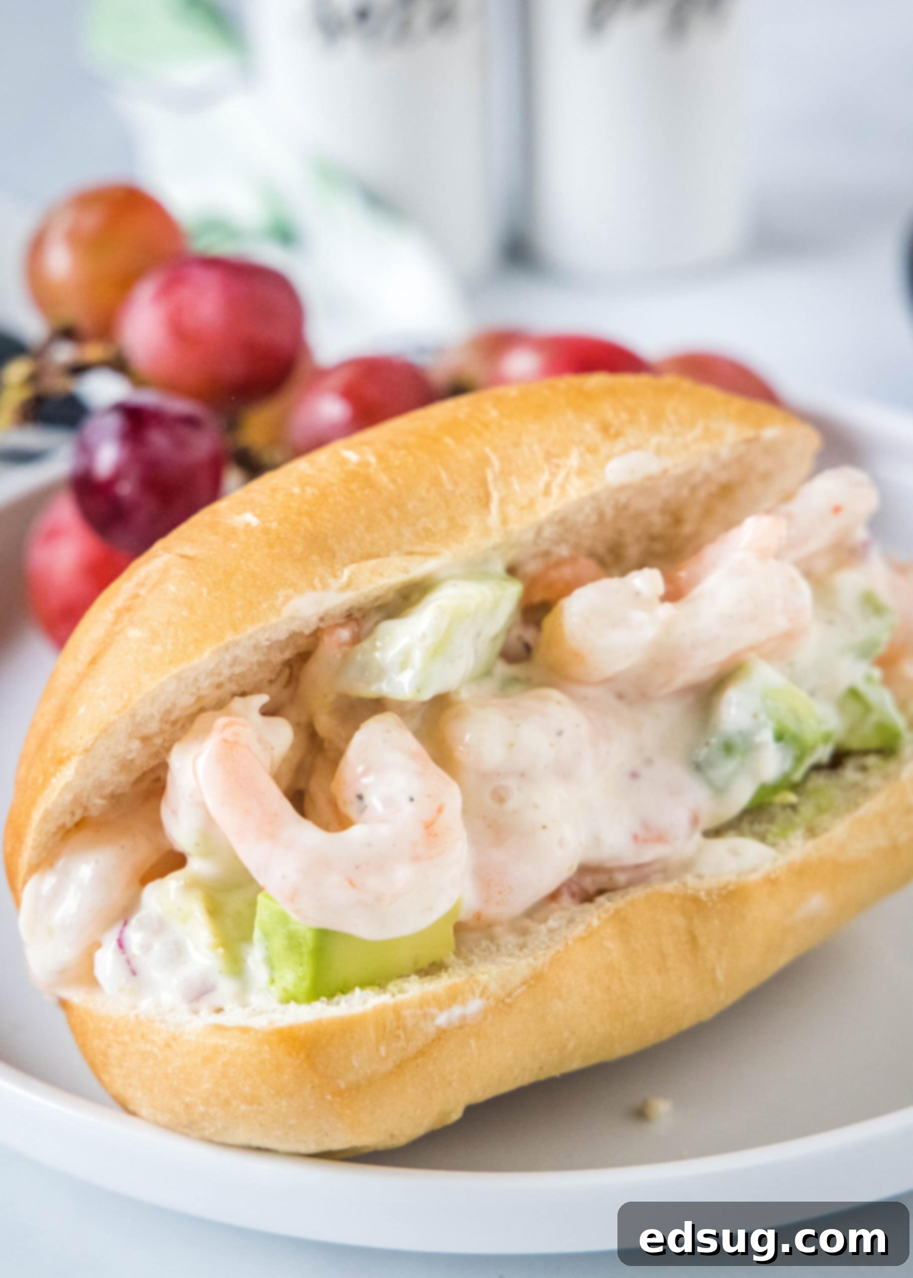 This creamy shrimp salad is about to become your favorite summer recipe! Made with chilled shrimp, crisp red onion, and Old Bay seasoning. A shrimp salad sandwich served on a white plate with red grapes in the background, showcasing its creamy texture.