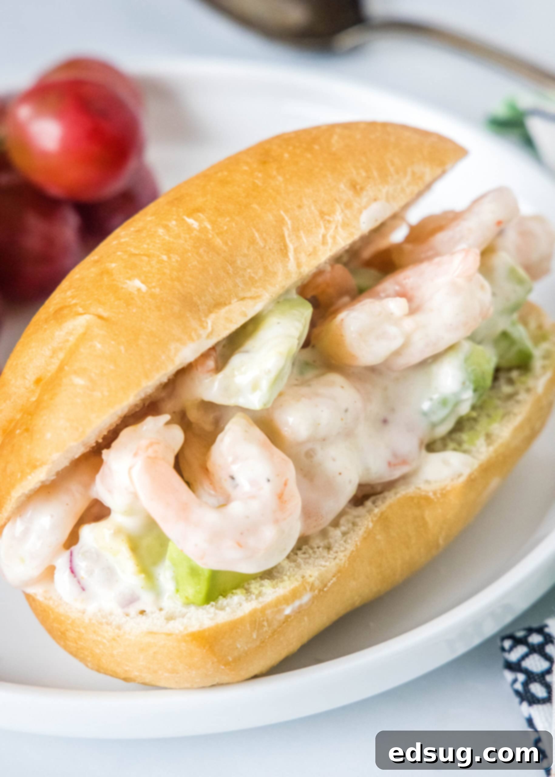 This creamy shrimp salad is about to become your favorite summer recipe! Made with chilled shrimp, crisp red onion, and Old Bay seasoning. A perfectly assembled shrimp salad sandwich served on a white plate with a side of fresh red grapes, ready for a delicious meal.