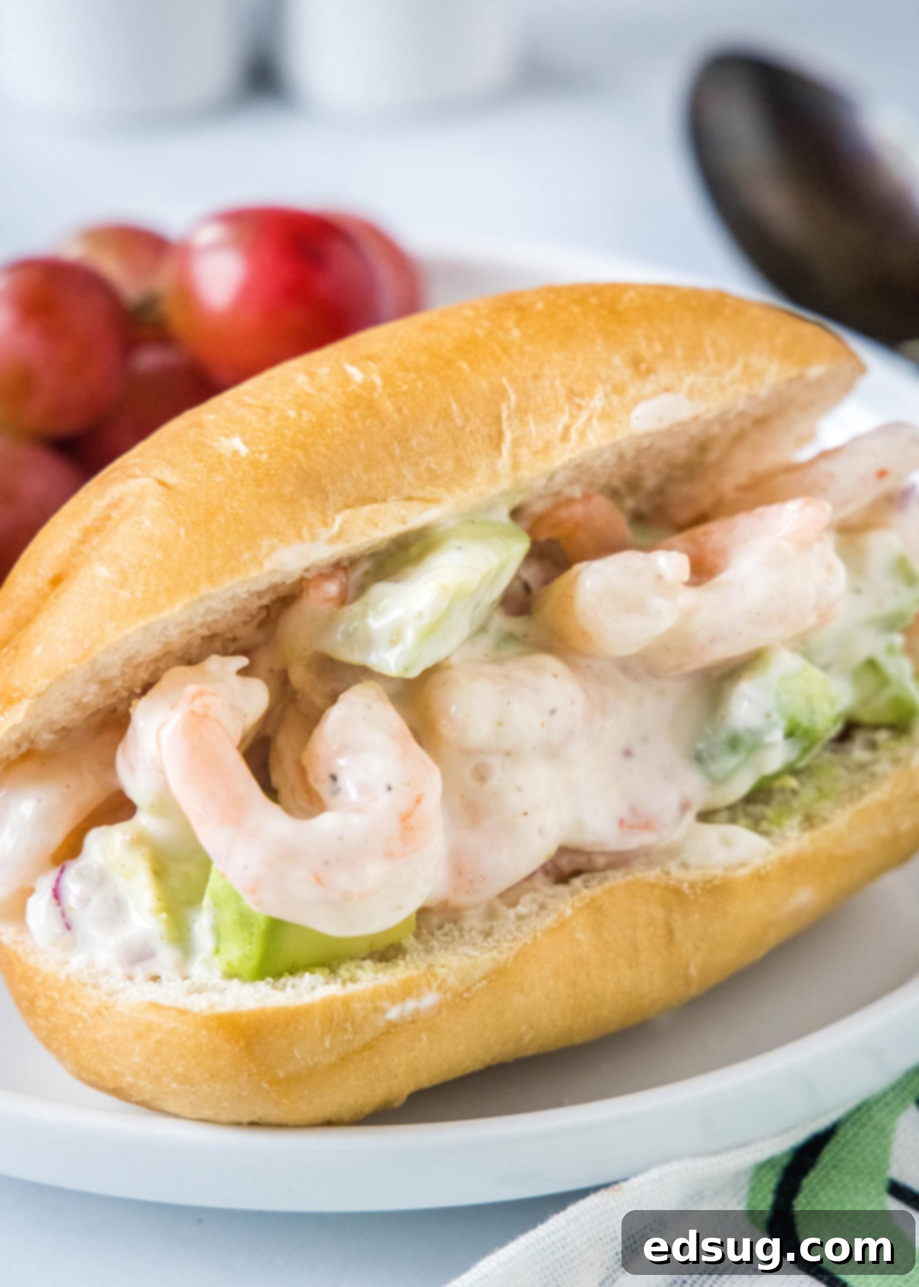 This creamy shrimp salad is about to become your favorite summer recipe! Made with chilled shrimp, crisp red onion, and Old Bay seasoning. Another view of a shrimp salad sandwich on a white plate with red grapes, highlighting its fresh appeal.