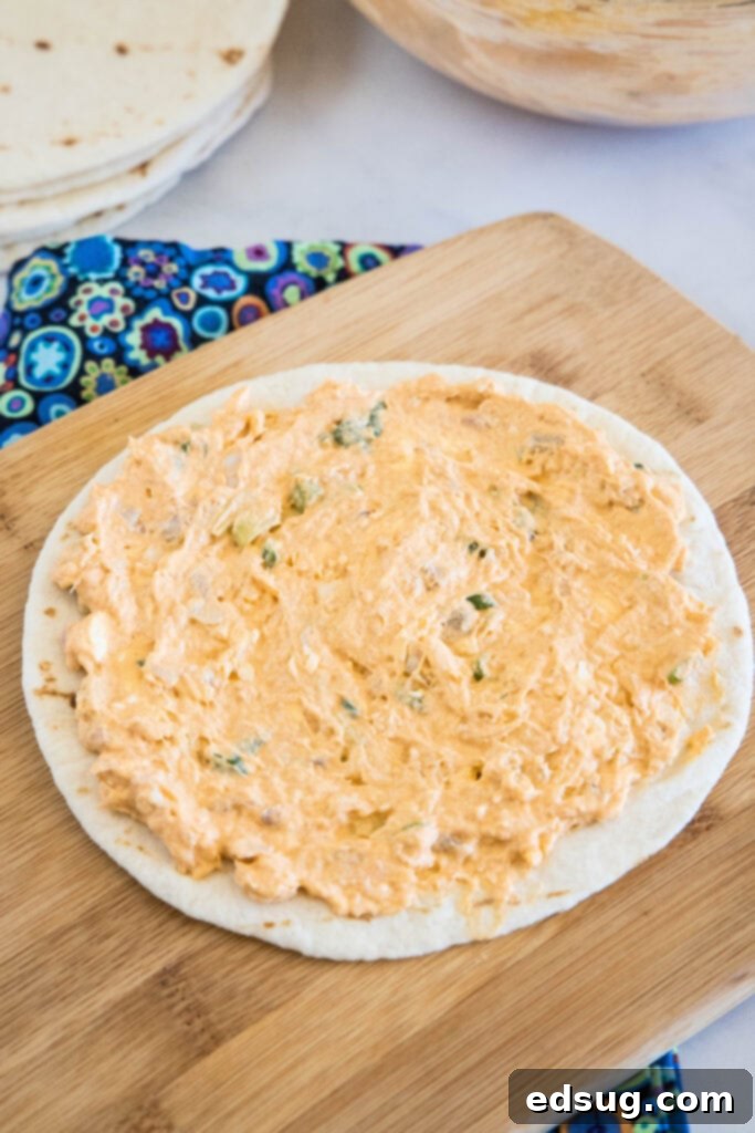 Buffalo chicken pinwheels are a quick and easy appetizer packed with bold Buffalo flavor. Roll up the spicy chicken filling, chill, and slice! The savory Buffalo chicken filling meticulously spread across a soft flour tortilla, laid out on a rustic wooden cutting board, ready for rolling.