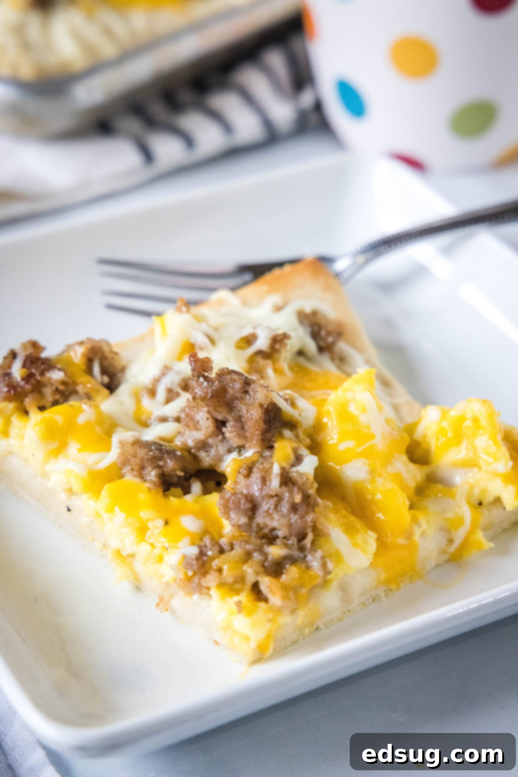 This satisfying breakfast pizza combines all of your breakfast favorites on top of a crispy pizza crust. It's so easy, baked in a sheet pan! A slice of breakfast pizza on a white plate next to a fork, showcasing its golden crust and delicious toppings.