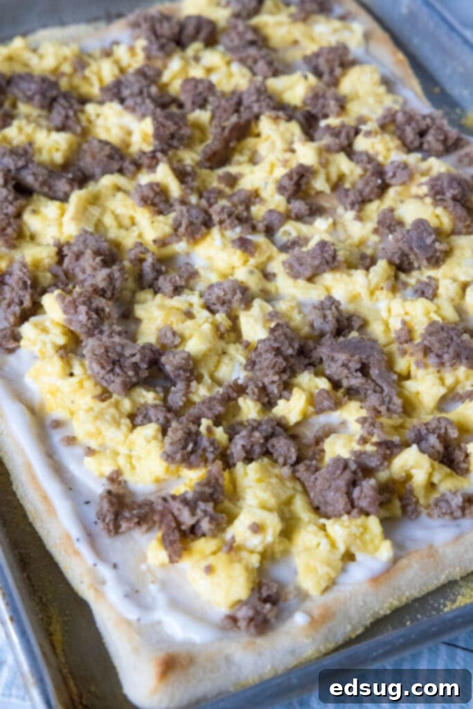 This satisfying breakfast pizza combines all of your breakfast favorites on top of a crispy pizza crust. It's so easy, baked in a sheet pan! Breakfast pizza assembly: crust topped with gravy, cooked sausage, and scrambled eggs, ready for cheese.