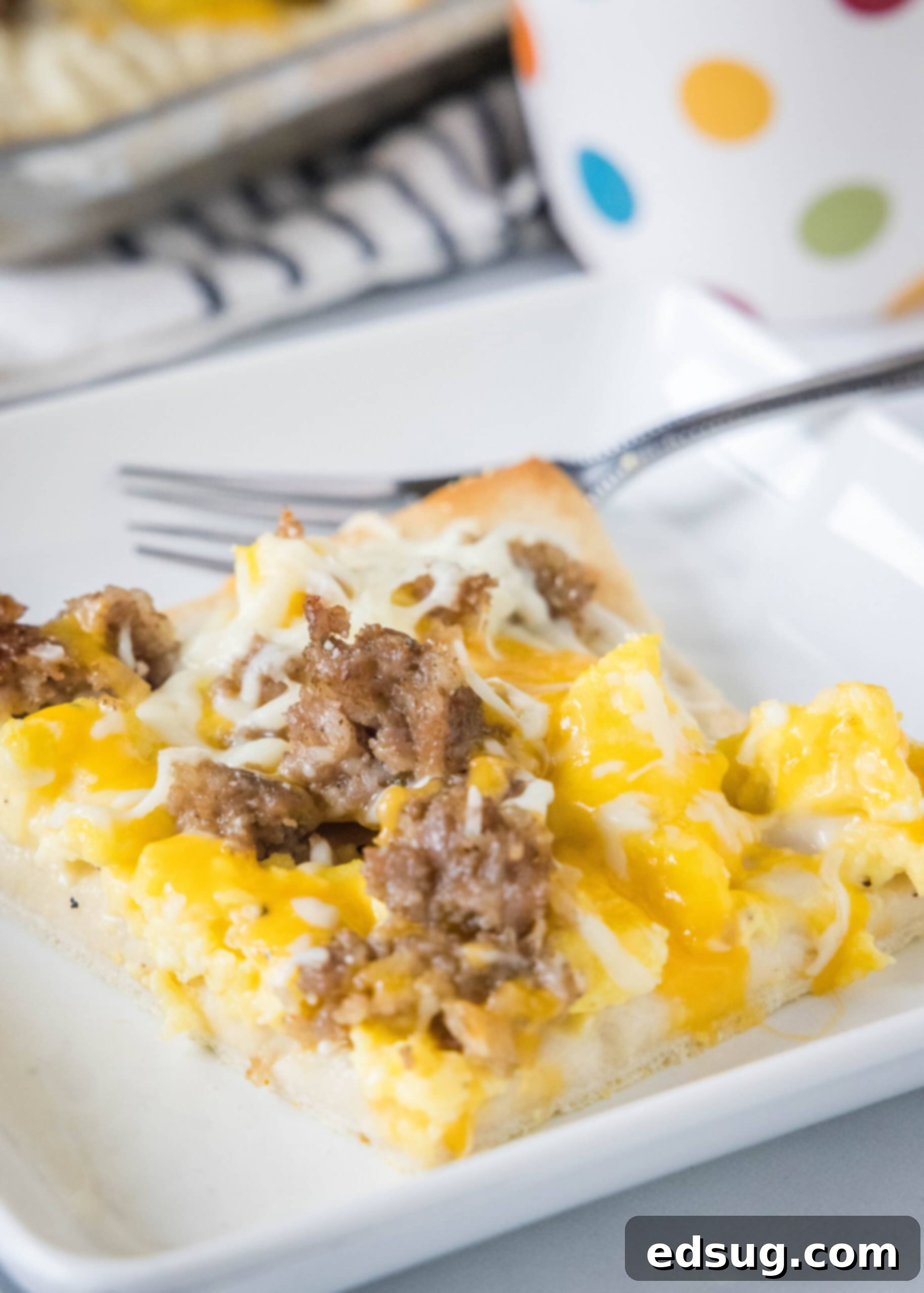 This satisfying breakfast pizza combines all of your breakfast favorites on top of a crispy pizza crust. It's so easy, baked in a sheet pan! A perfectly cooked slice of breakfast pizza on a white plate next to a fork, ready to be enjoyed.