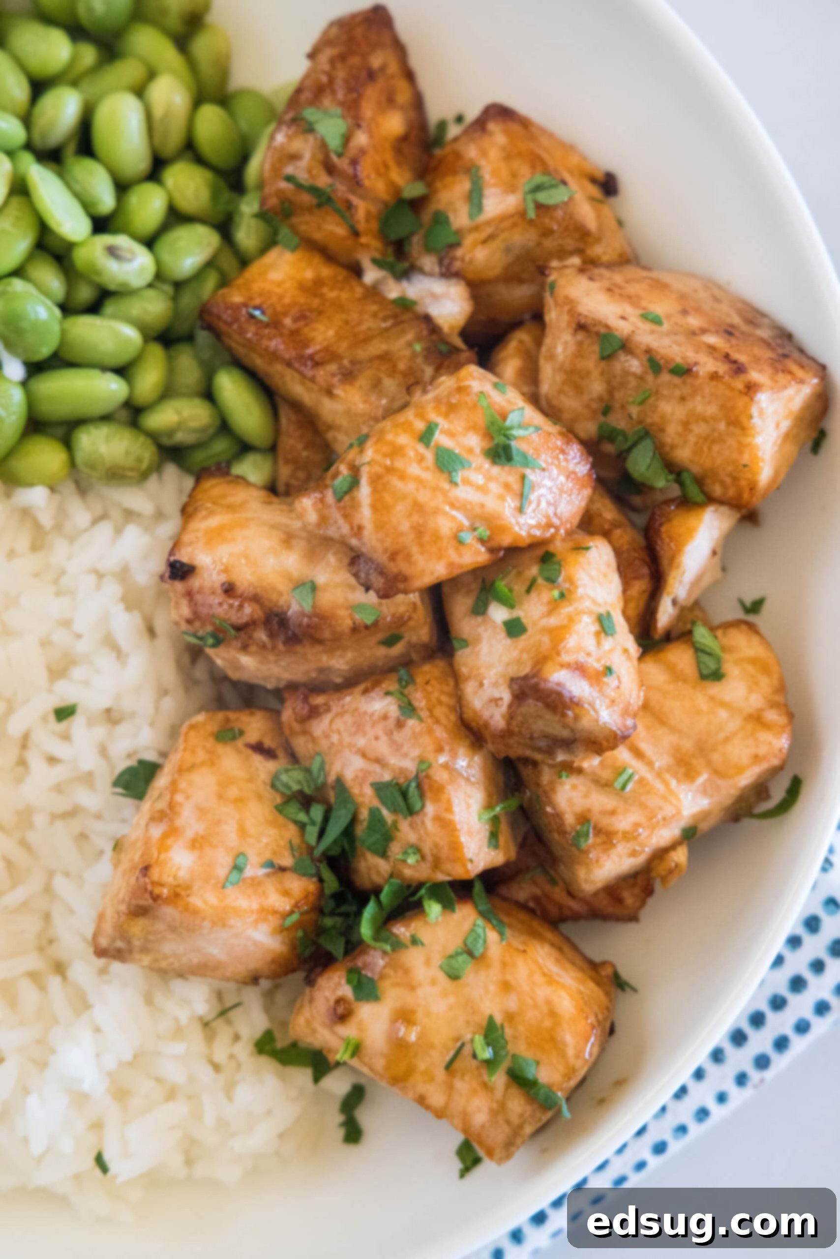 A close-up shot of perfectly cooked salmon bites, served alongside fluffy white rice and bright green edamame in a white bowl, ready to be enjoyed.