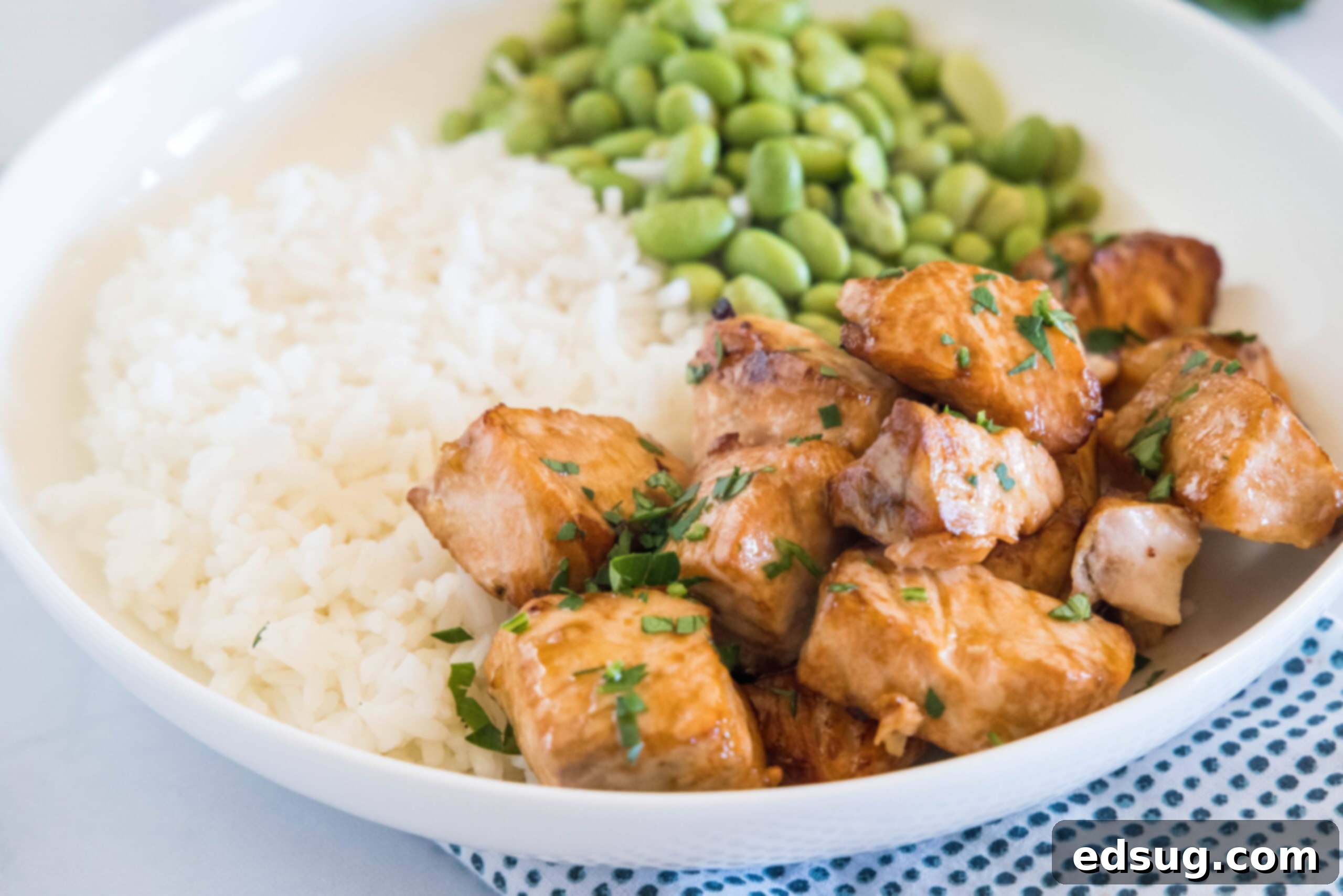 A beautifully composed white bowl featuring golden-brown salmon bites, white rice, and vibrant edamame beans, ready to be enjoyed.