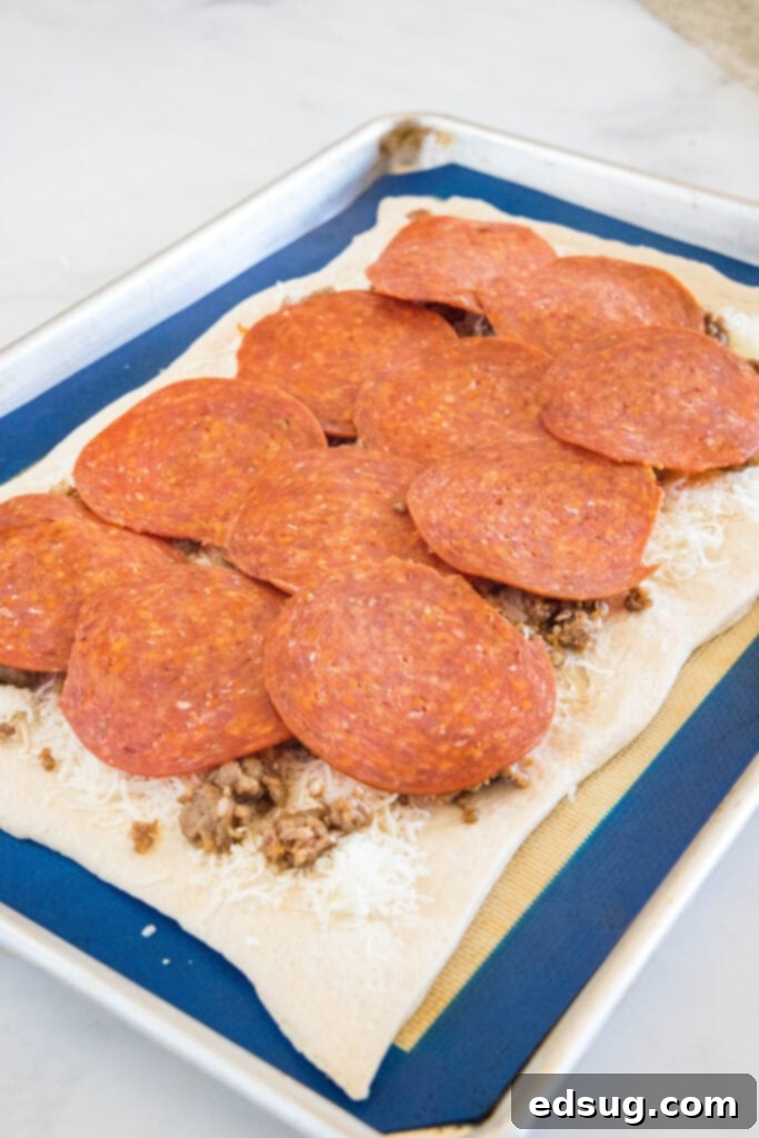 Put a fun spin on pizza night with this easy pizza bread recipe. Roll your favorite pizza toppings inside a pizza crust, bake, and slice! #pizzabread #pizza Pizza dough topped with shredded cheese, ground meat, and pepperoni slices on a baking sheet.