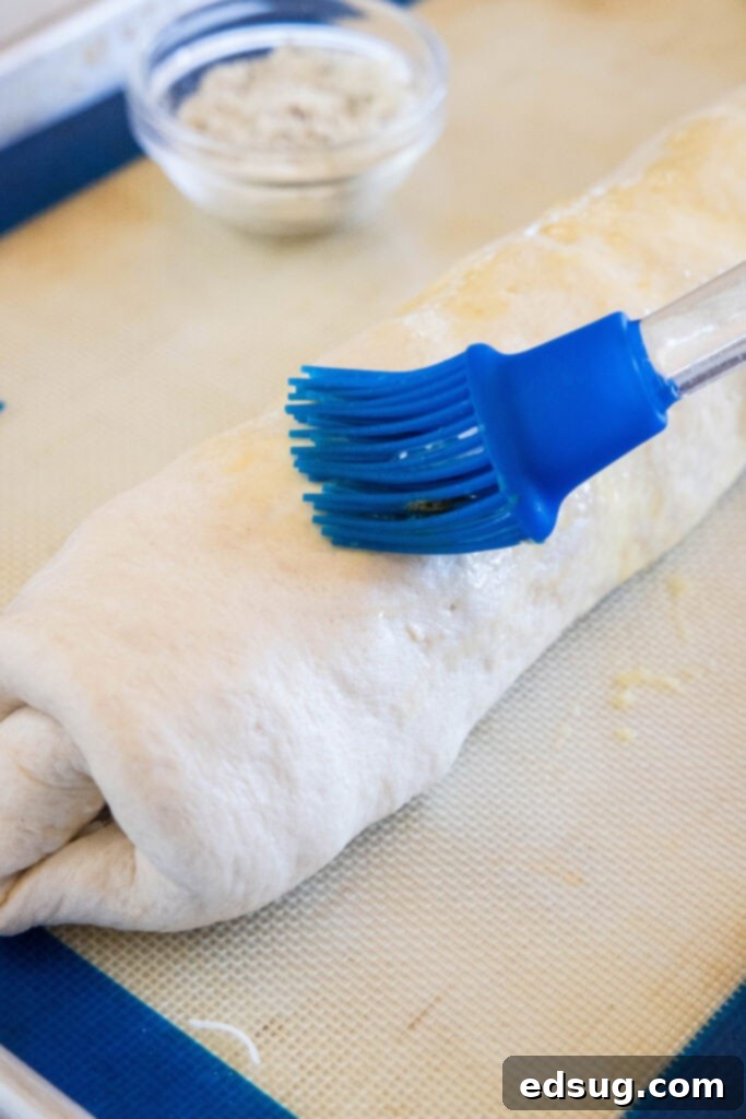 Put a fun spin on pizza night with this easy pizza bread recipe. Roll your favorite pizza toppings inside a pizza crust, bake, and slice! #pizzabread #pizza A blue basting brush brushing an egg wash over the top of rolled up pizza dough on a baking sheet.