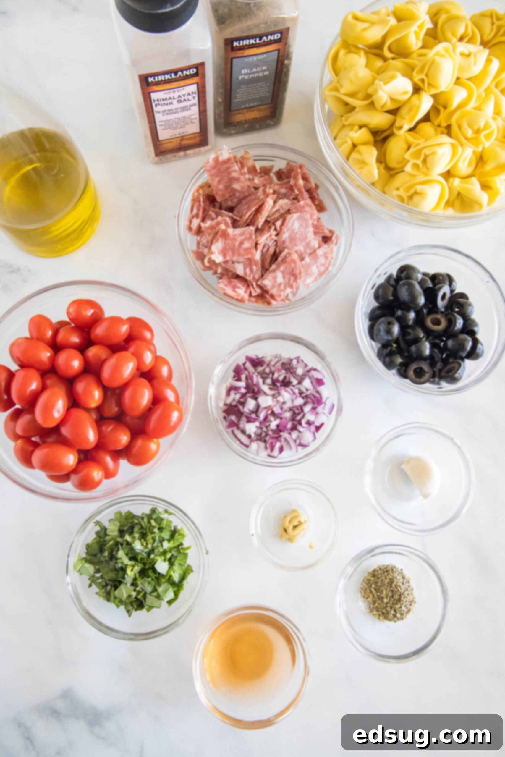 A collection of fresh ingredients laid out on a clean surface, including tortellini, tomatoes, olives, salami, and herbs, ready to be prepared for the pasta salad.
