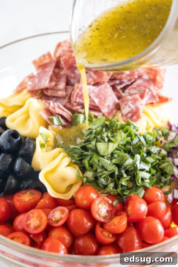 A stream of zesty Italian dressing being poured over the colorful tortellini salad ingredients in a clear glass bowl, ensuring every piece is coated with flavor.