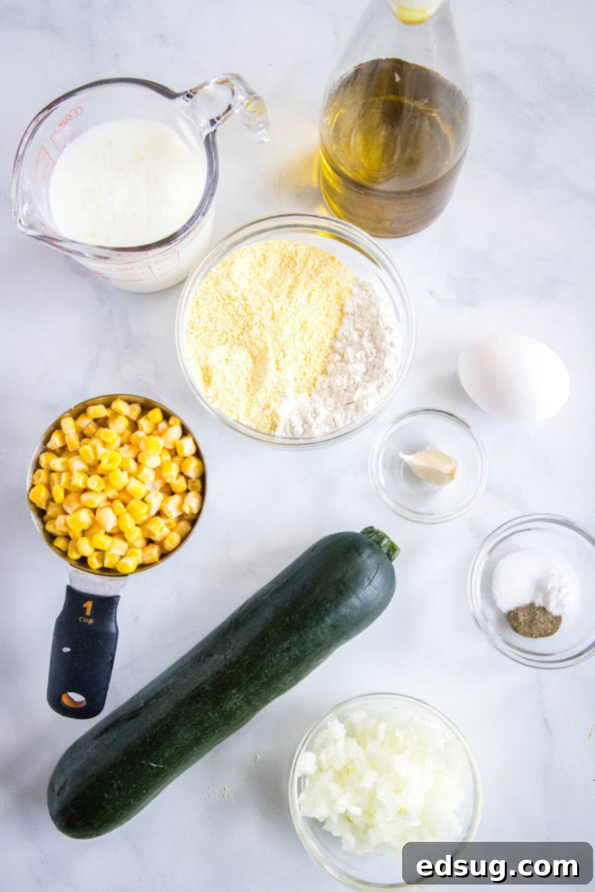 These easy zucchini fritters with corn are light, crispy, moist, and only take 25 minutes to make. They're a perfect side dish or snack! Overhead view of the fresh ingredients required for preparing zucchini and corn fritters: A vibrant whole zucchini, a measuring cup brimming with sweet corn kernels, a clear Pyrex dish holding creamy buttermilk, a bowl of finely chopped onions, a small bowl of cornmeal for texture, a single egg acting as a binder, a bowl of minced garlic for aromatic depth, and a separate bowl containing the seasoning blend of salt, black pepper, and a pinch of baking soda.