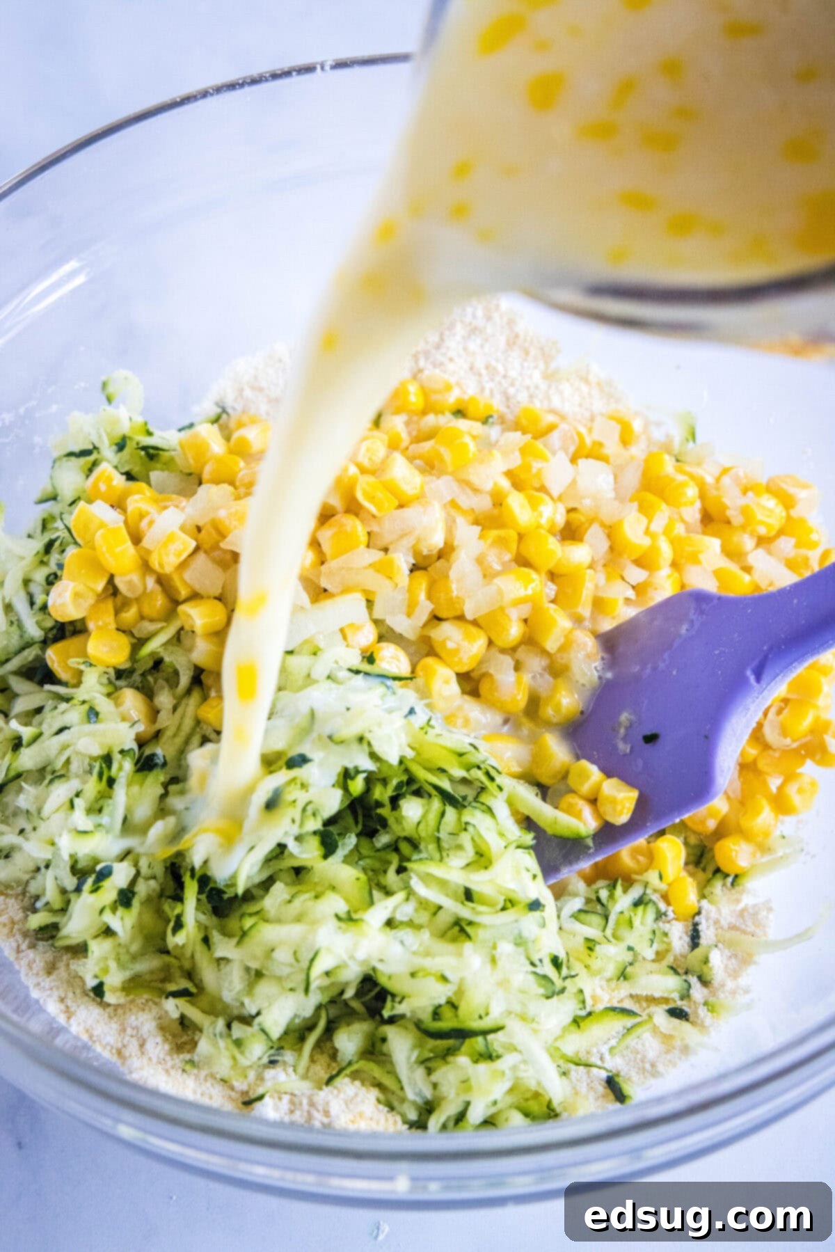 These easy zucchini fritters with corn are light, crispy, moist, and only take 25 minutes to make. They're a perfect side dish or snack! Buttermilk being poured into a mixing bowl with corn and shredded zucchini, with a spatula ready to mix, illustrating the final combining step of the fritter batter.