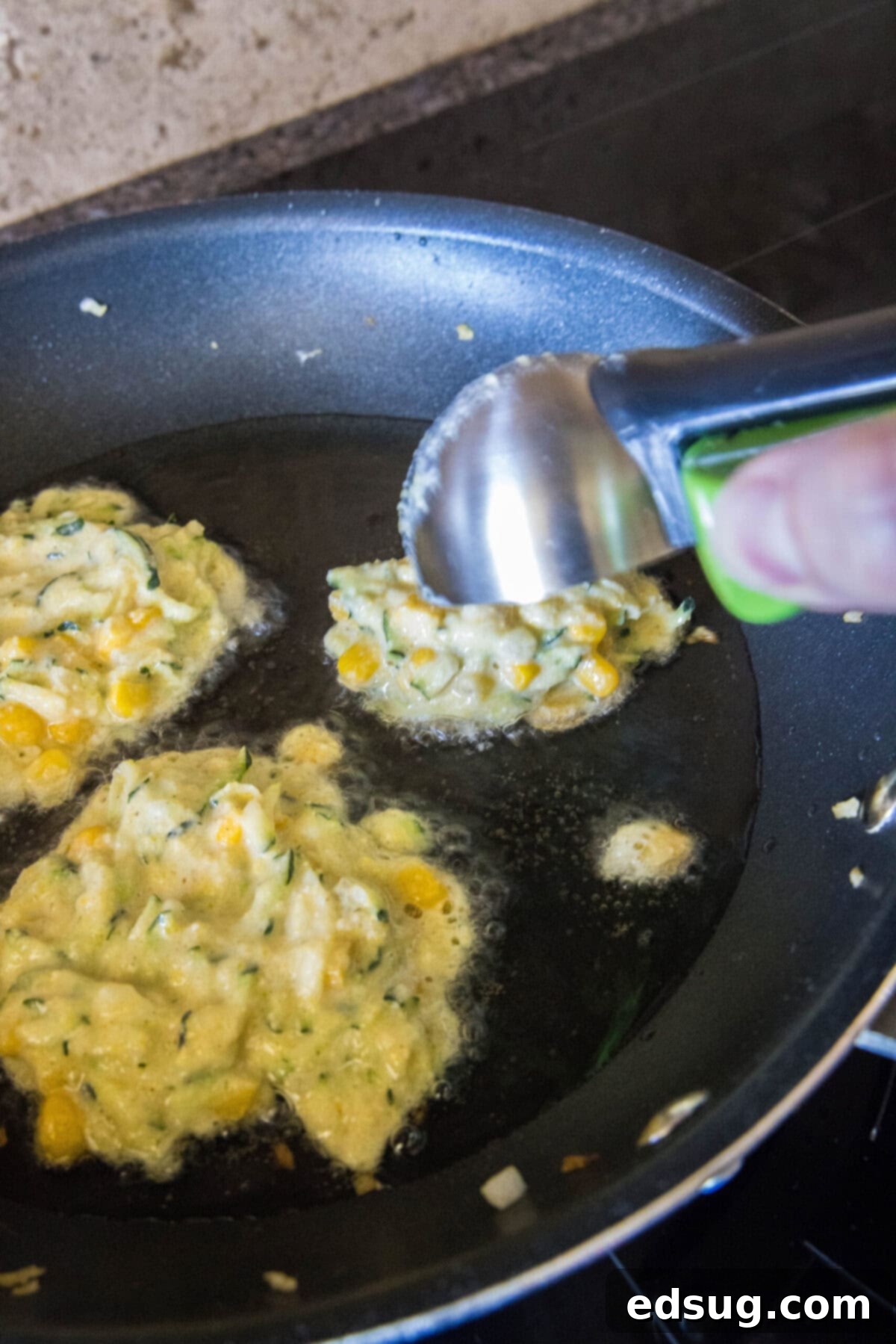 These easy zucchini fritters with corn are light, crispy, moist, and only take 25 minutes to make. They're a perfect side dish or snack! An ice cream scoop gently dropping a portion of zucchini fritter batter into a hot skillet, ready for frying.