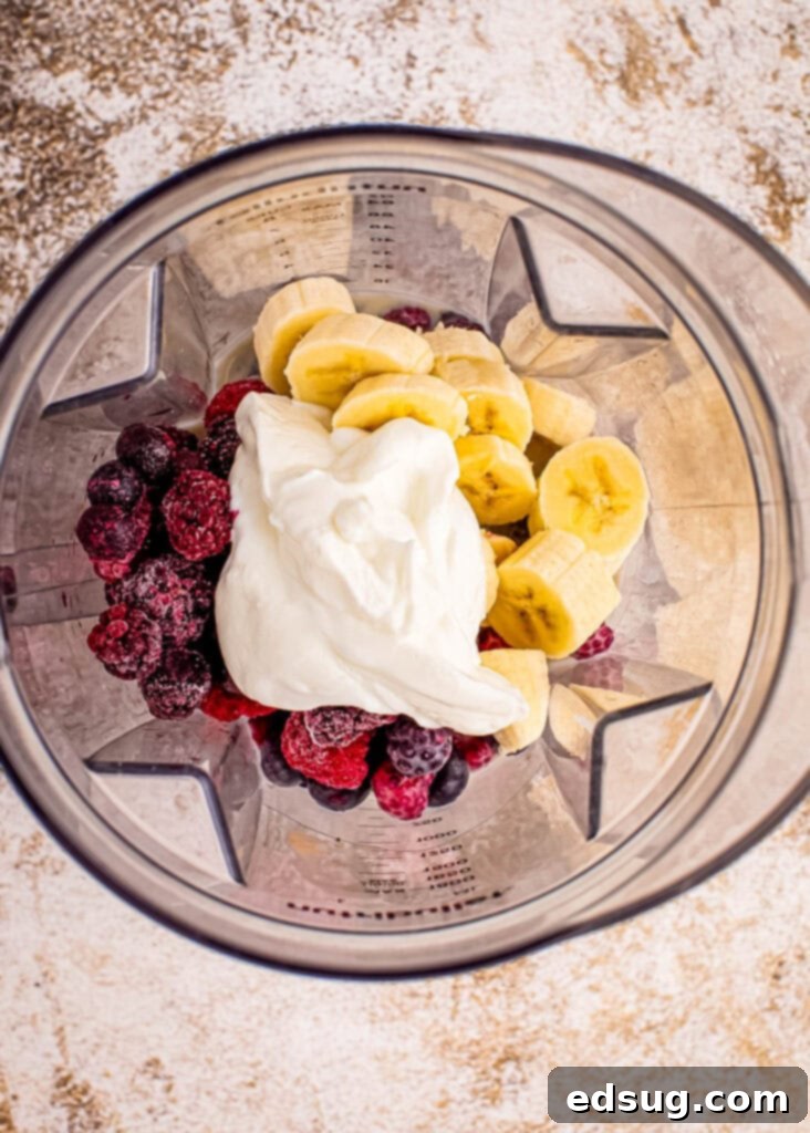 Frozen mixed berries, banana, milk, and Greek yogurt being added into a high-speed blender for a smoothie bowl