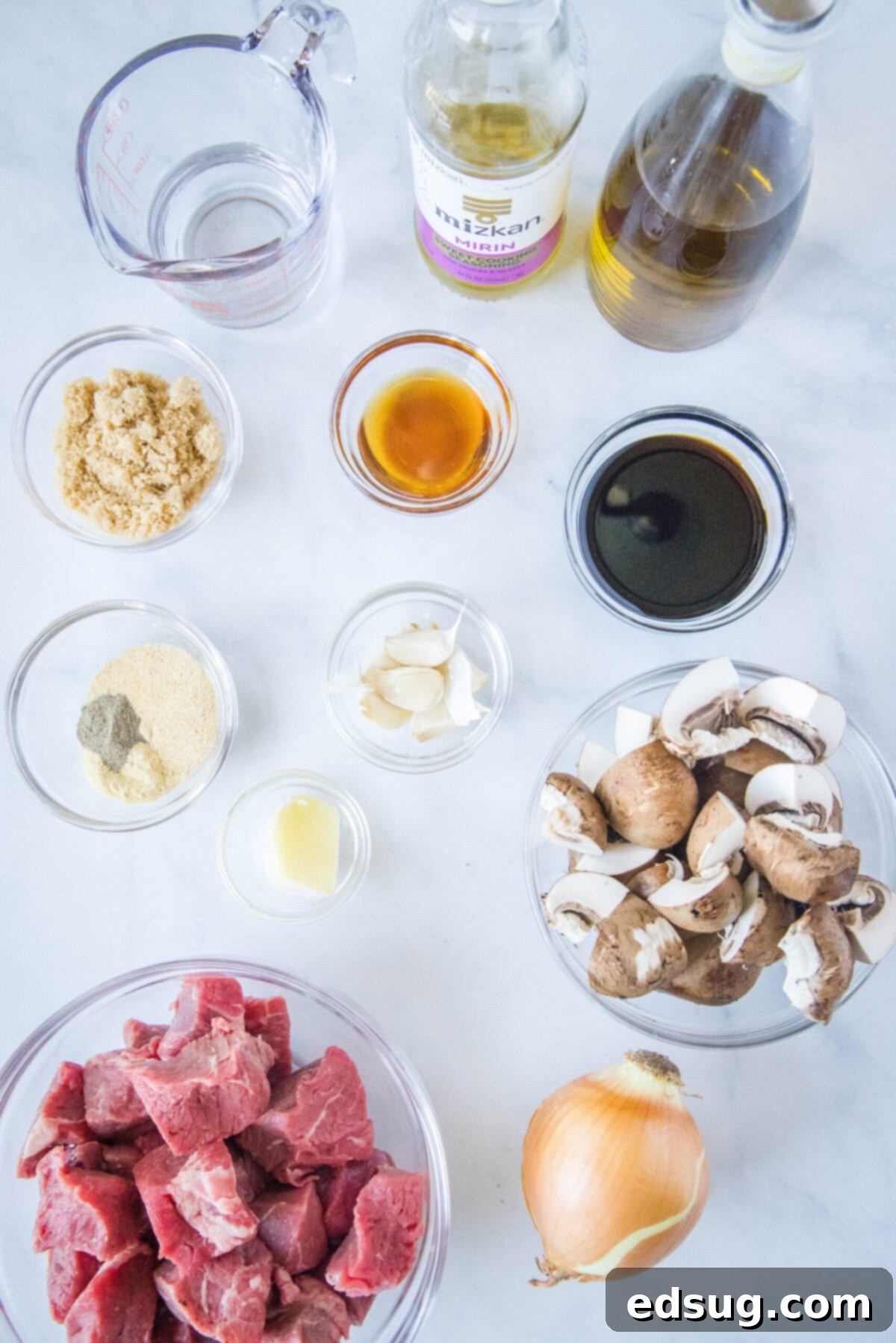 Overhead view of some of the ingredients needed for Korean beef kabobs: A bowl of beef cubes, a bowl of mushrooms, a bowl of garlic, a bowl of ginger, a bowl of brown sugar, a bowl of soy sauce, a bowl of sesame oil, a bowl of seasonings, a bottle of olive oil, a bottle of mirin, a pyrex of water, and a yellow onion