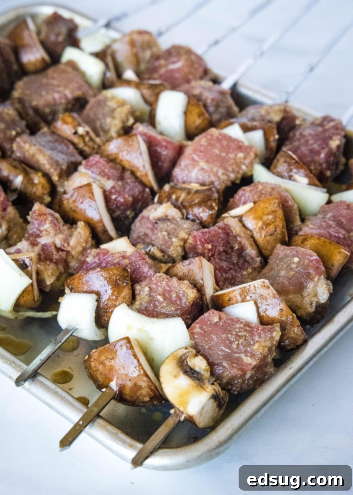 A baking sheet full of uncooked beef, onion, and mushroom kabobs, ready for the grill