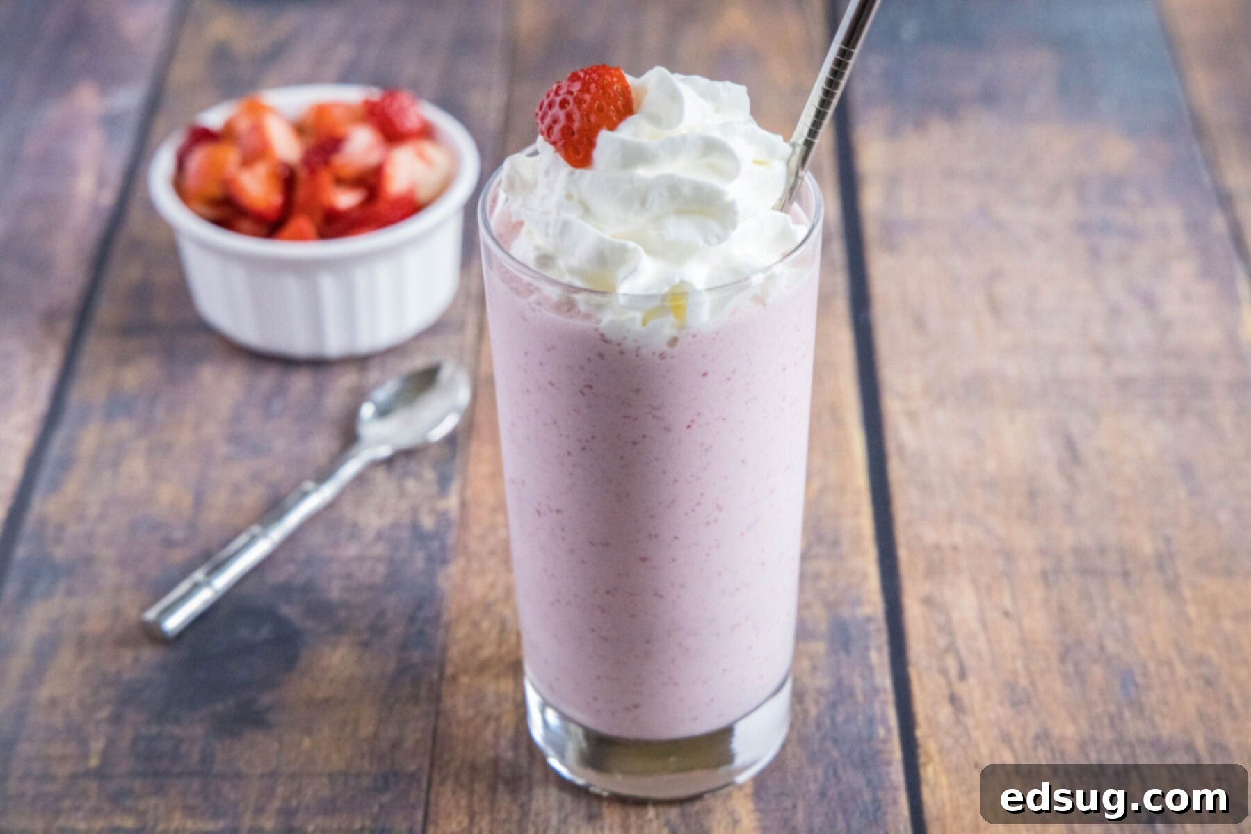 These 5-ingredient strawberry milkshakes are full of nostalgia and flavor. They're quick and easy to make, and absolutely delicious. Wide angle view of a strawberry milkshake topped with whipped cream, next to a spoon and a bowl of strawberries.