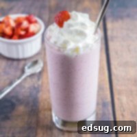 Wide angle view of a strawberry milkshake topped with whipped cream, next to a spoon and a bowl of strawberries.