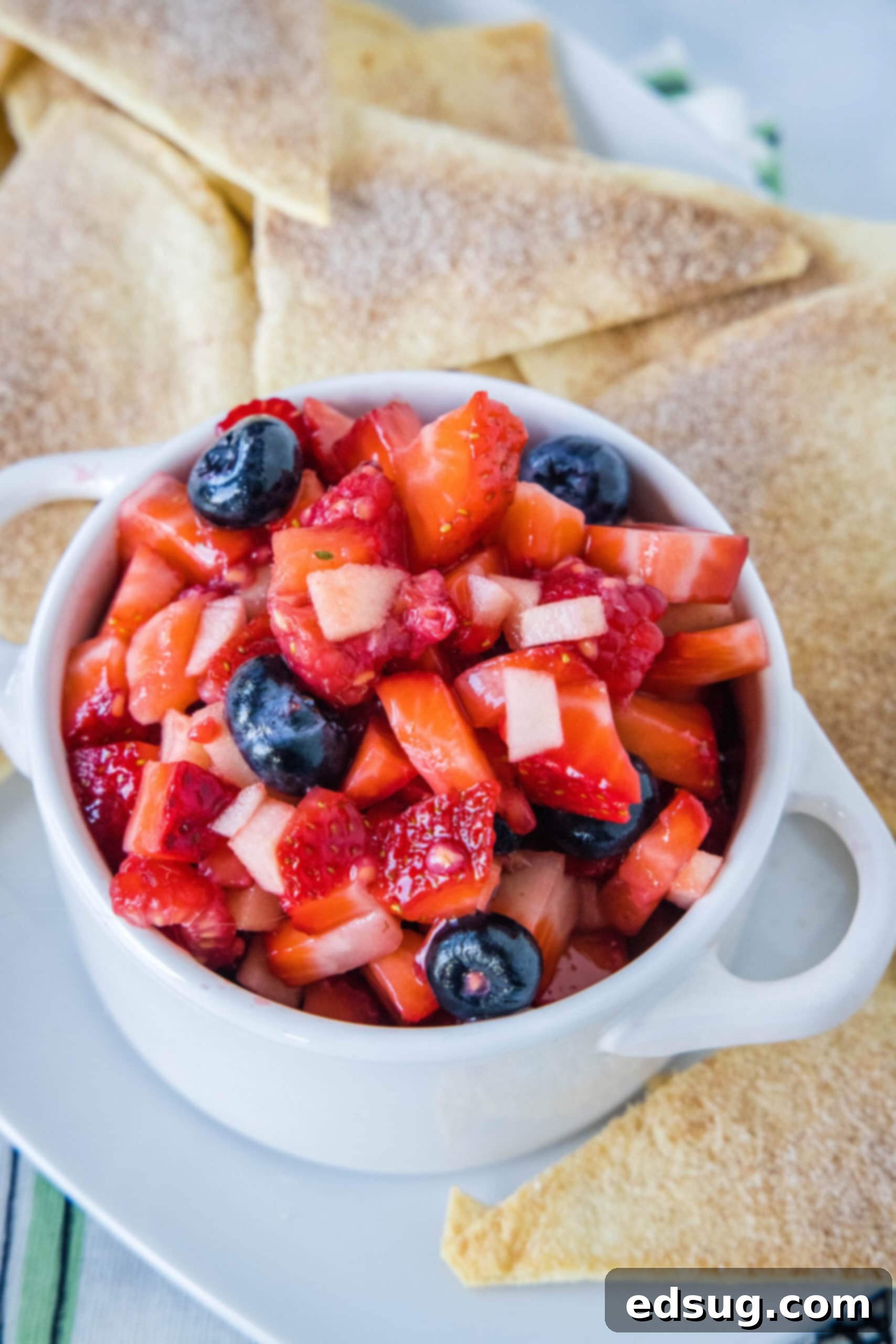 This fruit salsa is a sweet summer salsa recipe bursting with fresh seasonal berries. Serve it with cinnamon chips for a light and easy snack! Fruit salsa in a white ceramic serving bowl with two handles, surrounded by cinnamon chips.