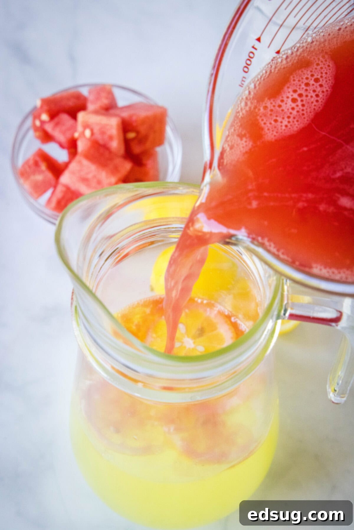 Sunny Watermelon Lemonade 4 A hand pouring vibrant red watermelon juice into a pitcher of yellow lemonade.