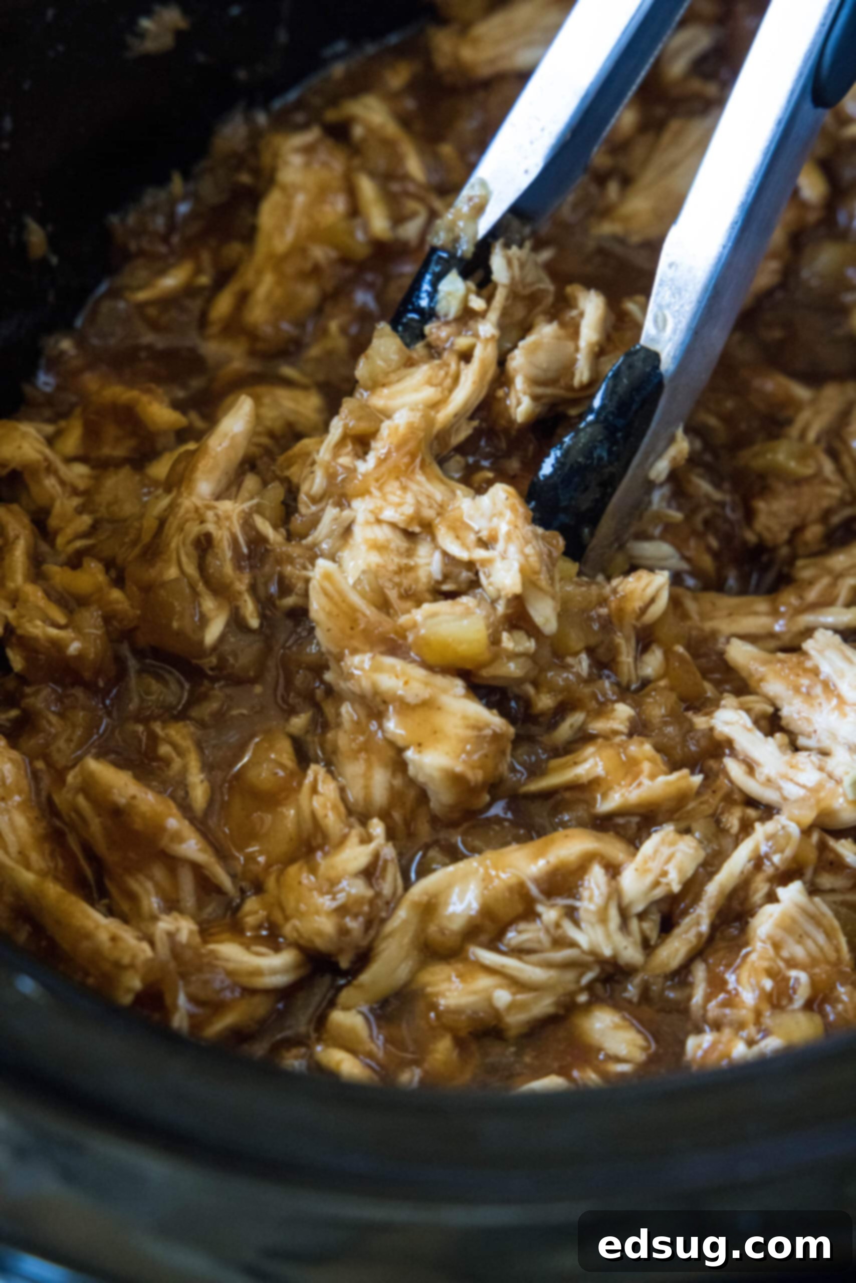 A pair of tongs lifting succulent shredded Hawaiian chicken from the slow cooker, glistening with its sweet and smoky sauce.
