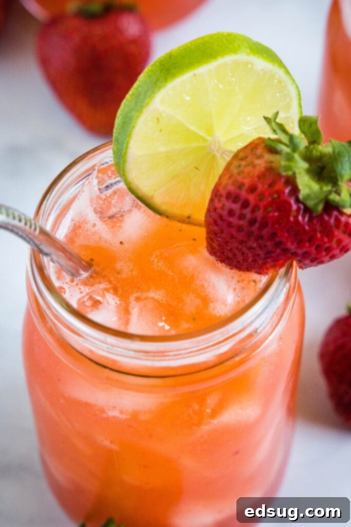 A close-up of a hand pouring vibrant red strawberry agua fresca from a clear glass pitcher into a glass filled with ice, garnished with a fresh lime slice.